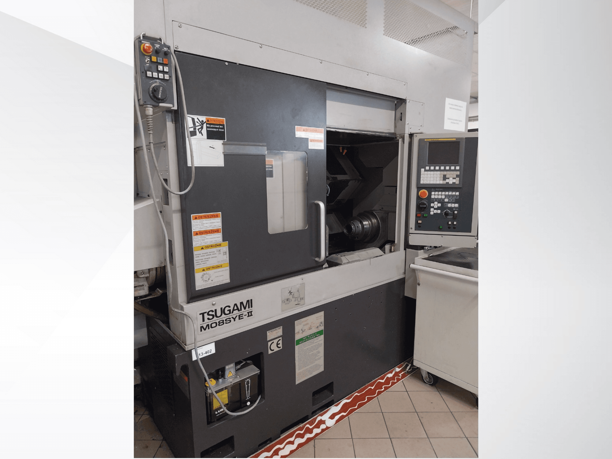 Tokarka CNC Tsugami M08SY-II, widok z przodu, pokazujący panel sterowania, wrzeciono i obszar załadunku. Zawiera uwagi dotyczące bezpieczeństwa i etykietę marki.