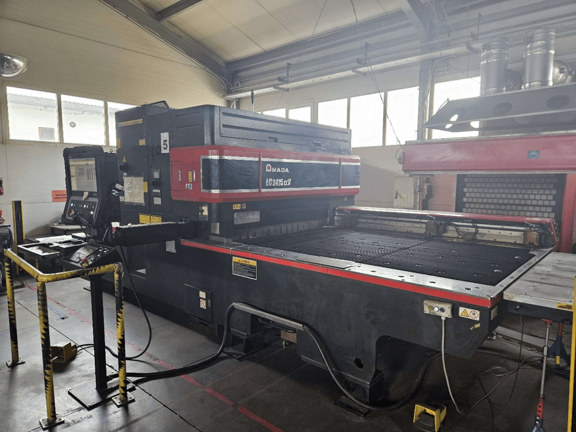 Maszyna do cięcia laserowego CO₂ AMADA LC2415 Alpha 5 – widok z przodu przedstawiający panel sterowania oraz stół roboczy z barierami zabezpieczającymi.