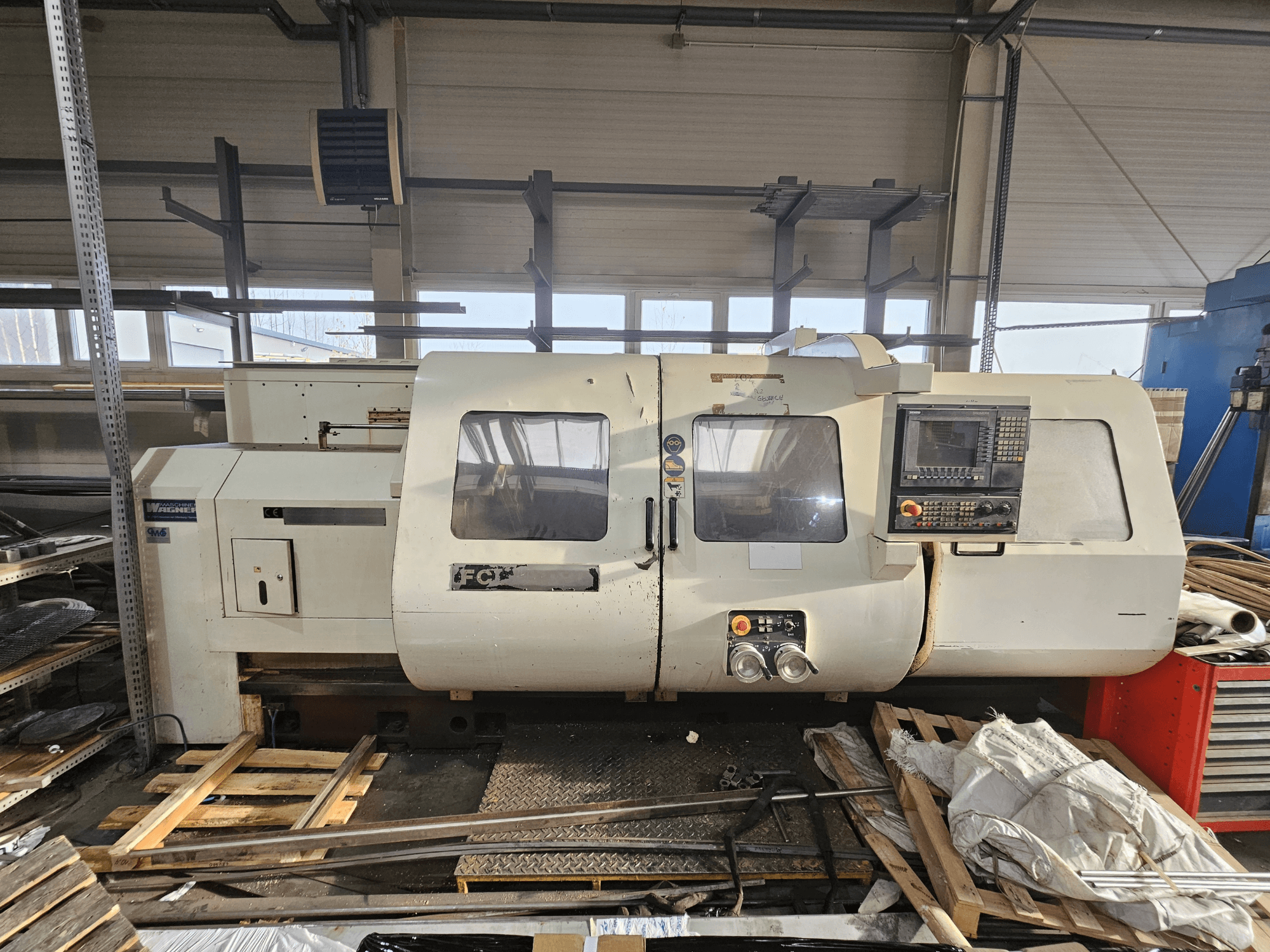 Tokarka CNC Wabeco, widok z boku, z panelem sterowania, oknami i obszarem roboczym; maszyny na drewnianych paletach w otoczeniu warsztatu.