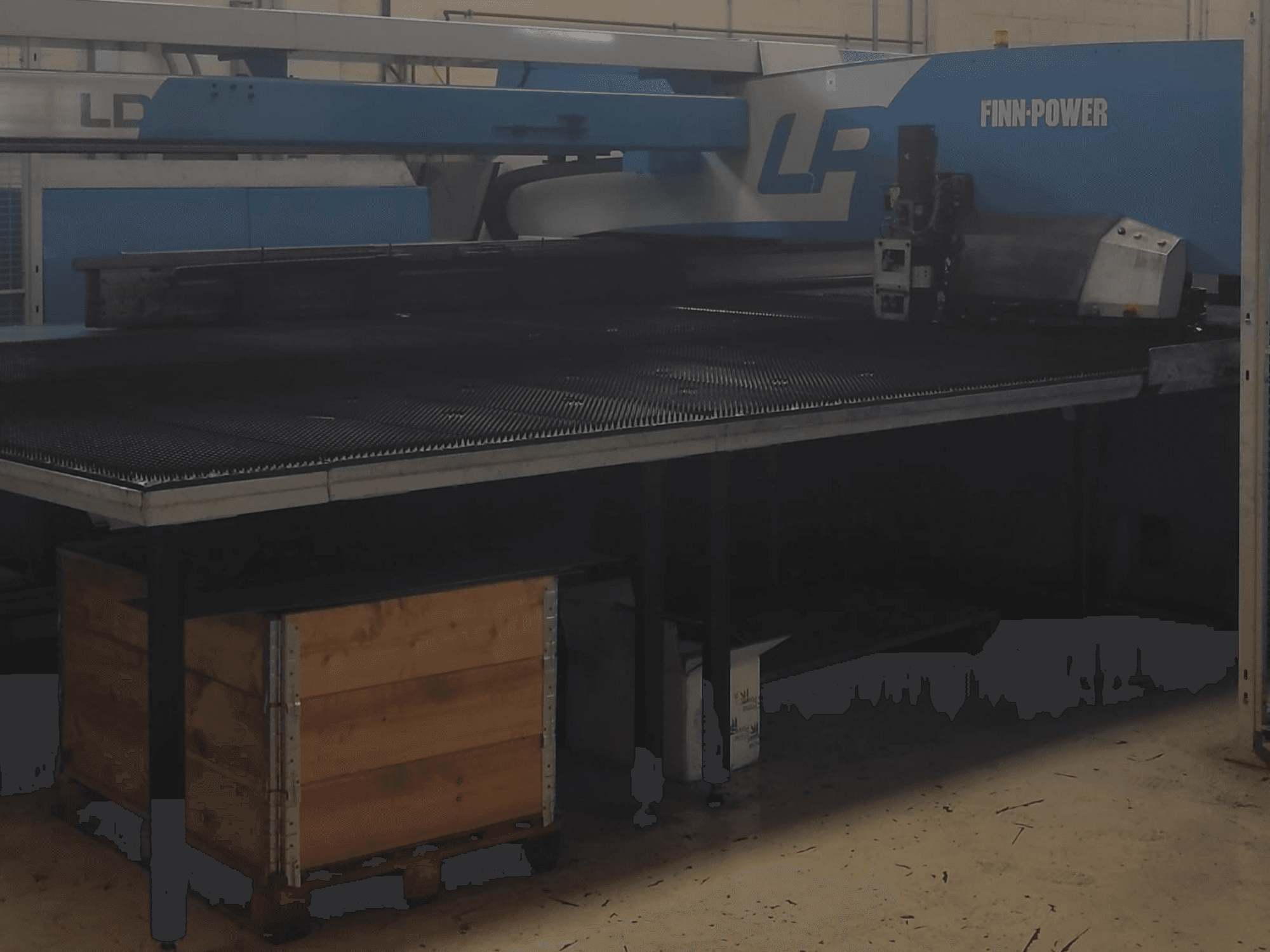 Maszyna do cięcia Finn-Power LP w fabryce, widok z przodu z teksturowanym metalowym stołem i drewnianą skrzynią pod spodem.