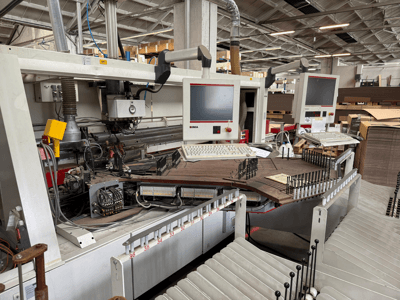 Precyzyjna maszyna do obróbki drewna IMA z dwoma monitorami i panelami sterowania, widok z przodu pokazujący przenośnik i obszar narzędzi.