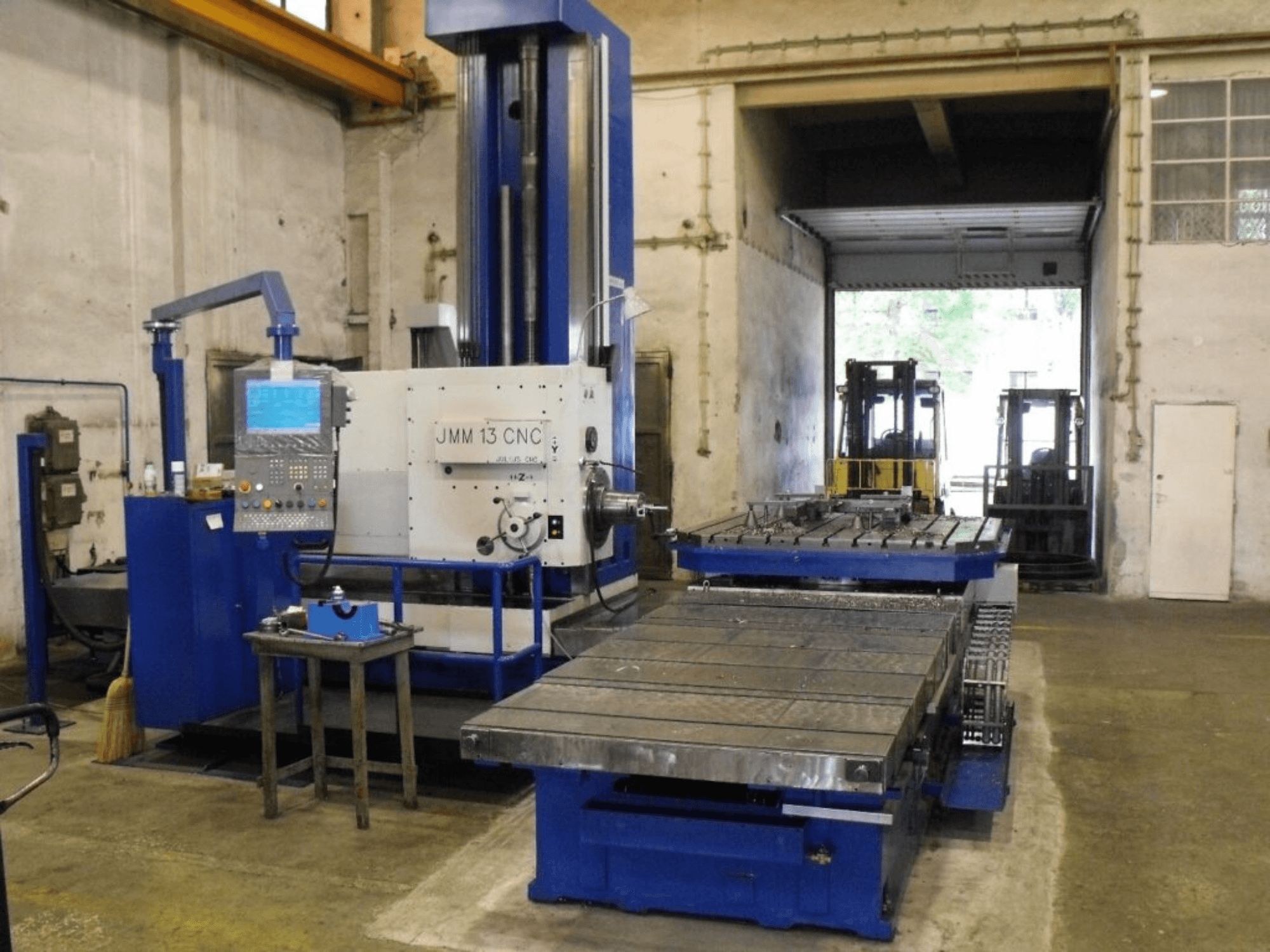 Frezarka CNC JMM 13 w warsztacie, widok z przodu, z panelem sterowania, dużym stołem roboczym i tłem przemysłowym.