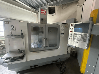 Maszyna CNC Haas VF-2, widok z przodu, z elementami sterującymi, uchwytem narzędziowym i przezroczystymi drzwiami z widocznym oprzyrządowaniem wewnątrz obudowy.