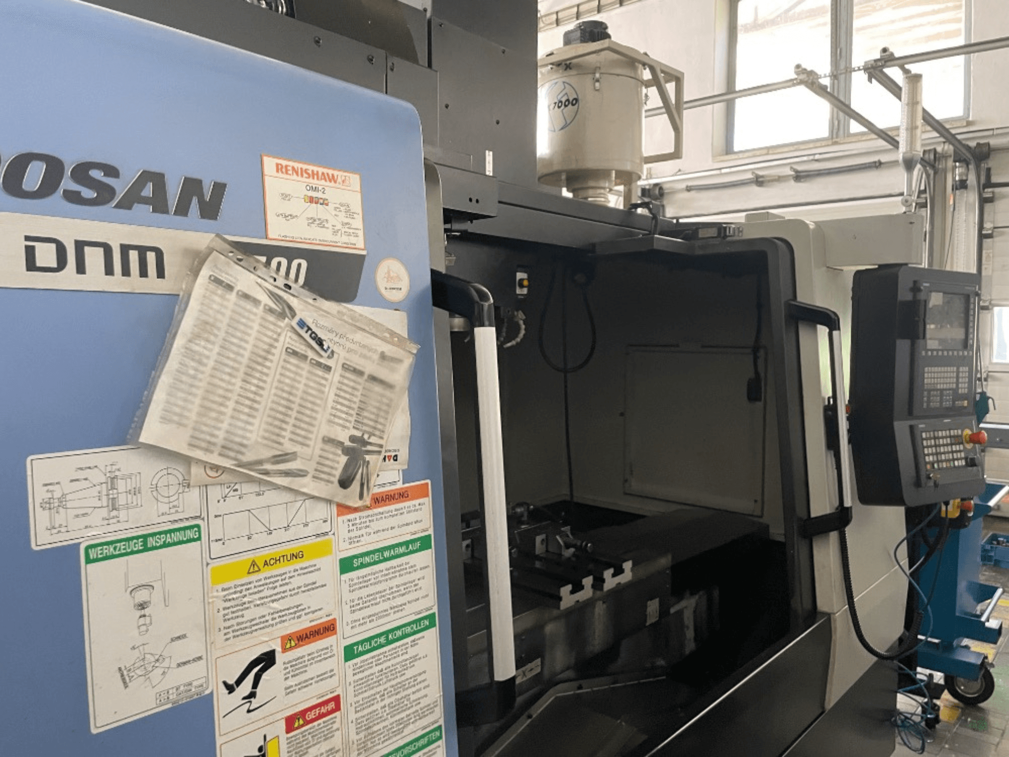 Pionowe centrum obróbcze DOOSAN DNM 5700, widok z przodu, pokazuje panel sterowania, etykiety bezpieczeństwa i szczegóły maszyny.