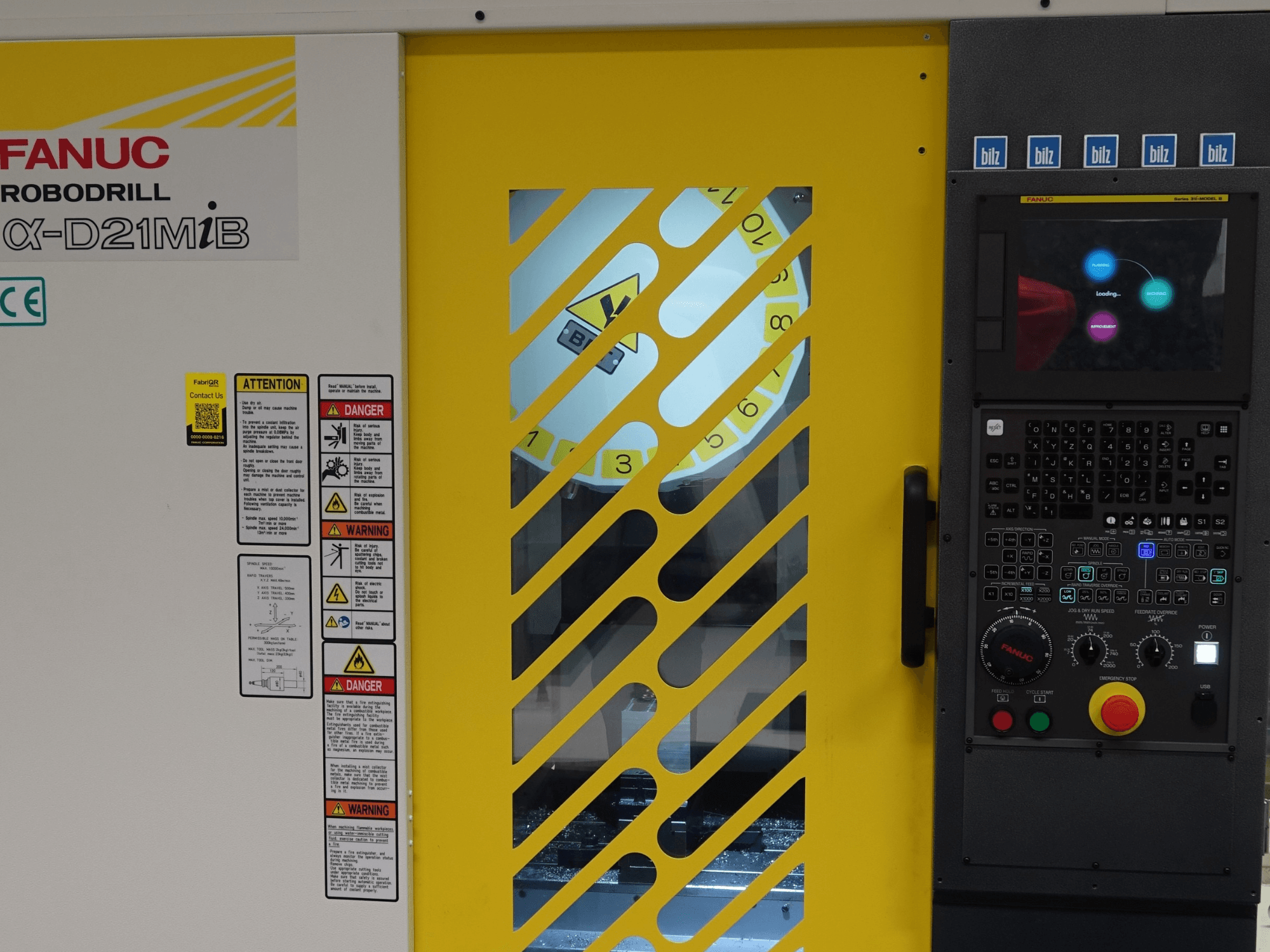 Widok z przodu pionowego centrum obróbczego FANUC α-D21MiB, na którym widoczne są ostrzeżenia bezpieczeństwa, panel sterowania oraz żółte drzwi bezpieczeństwa.