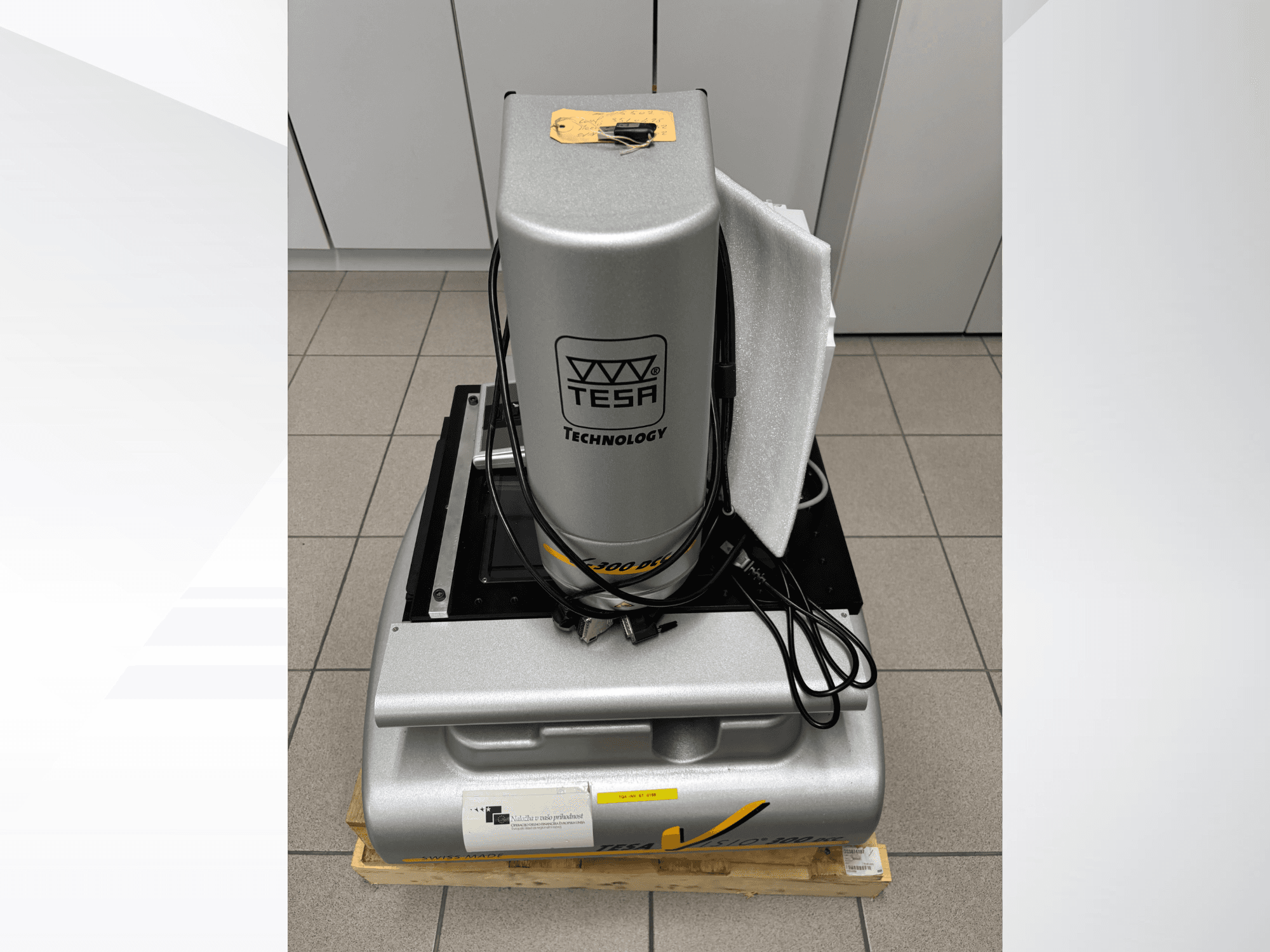 Optyczna maszyna pomiarowa TESA Visio 300 DCC w widoku z przodu z kablami, na drewnianej palecie i widocznym opakowaniem ochronnym.