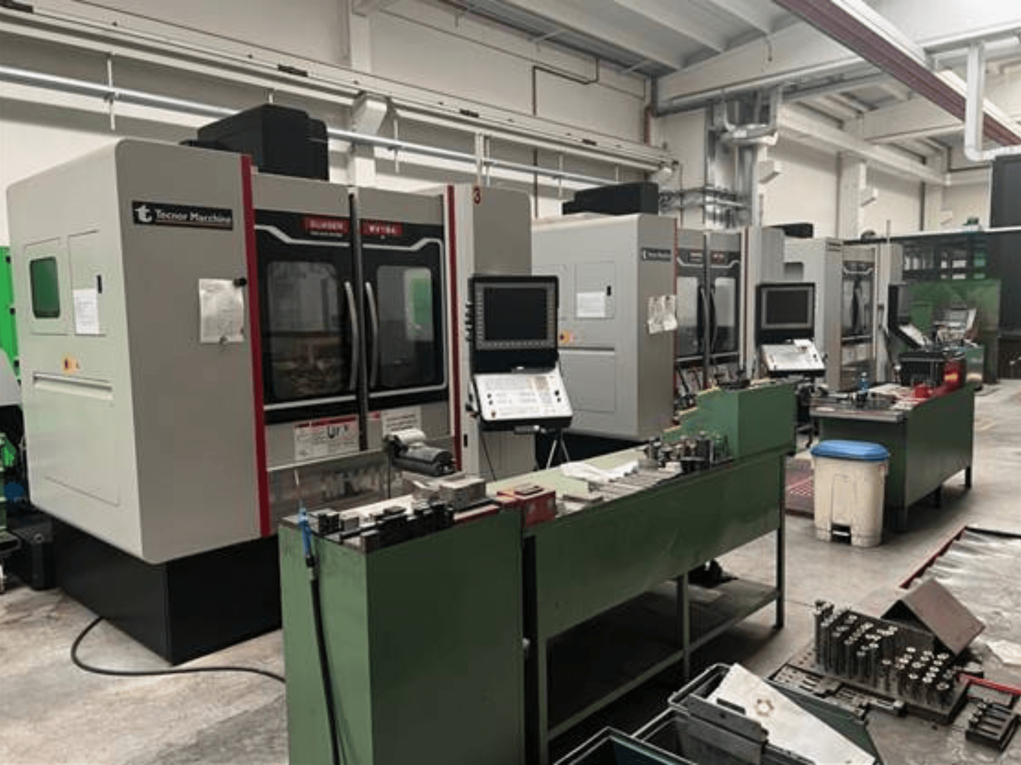 Cztery maszyny CNC firmy Tecnoer Marchine w warsztacie, widok z przodu, z narzędziami i częściami na zielonych stołach warsztatowych.