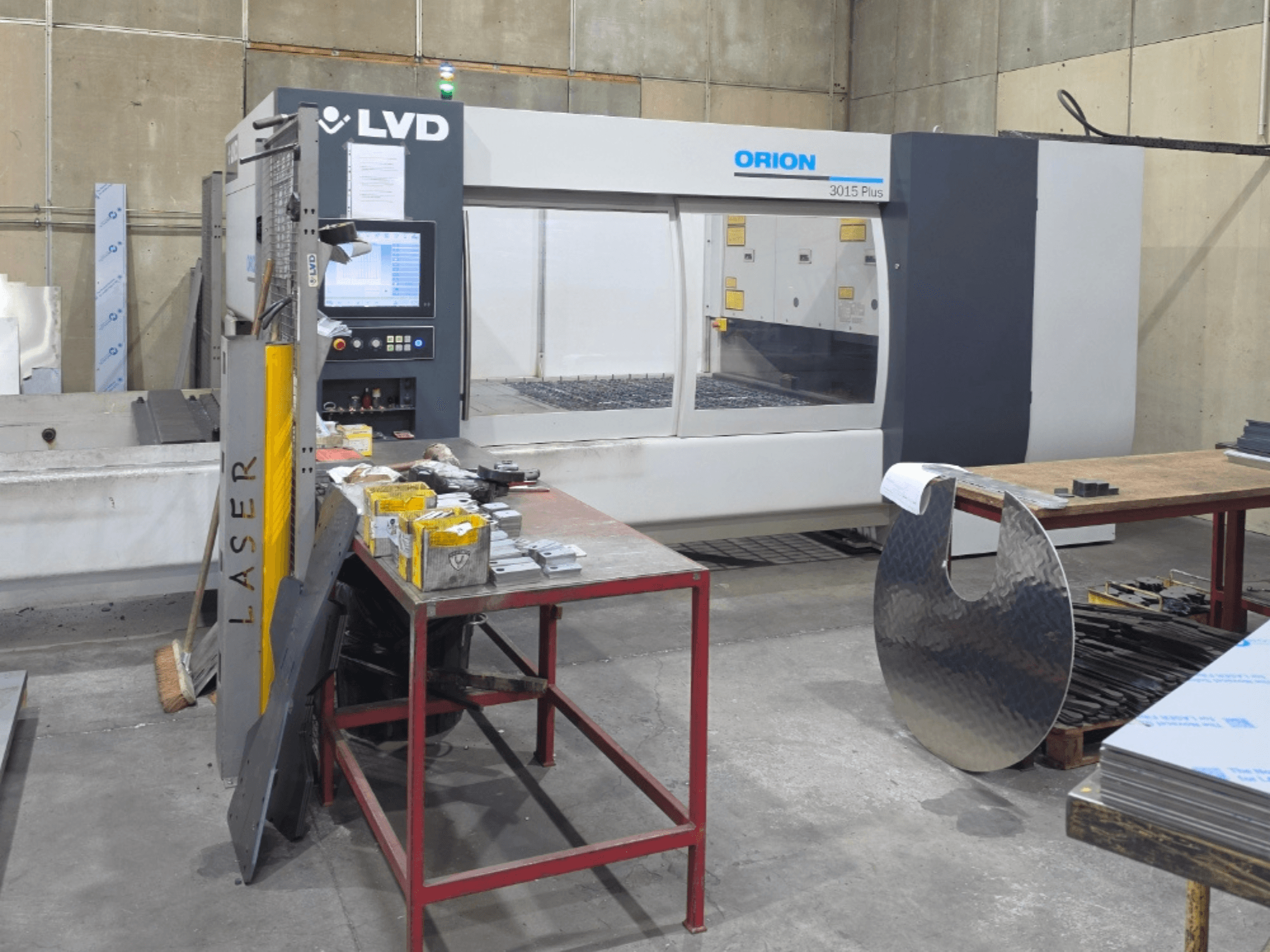 Maszyna do cięcia laserowego LVD Orion 3015 Plus z laserem CO₂, widok z przodu z panelem sterowania; otoczona metalowymi elementami na stole roboczym.