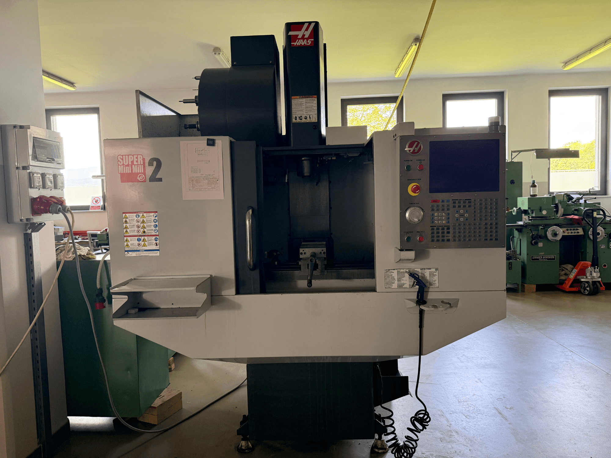 Pionowe centrum obróbcze Haas Mini Mill 2 w widoku z przodu, przedstawiające panel sterowania i obszar narzędzi z etykietami ostrzegawczymi.
