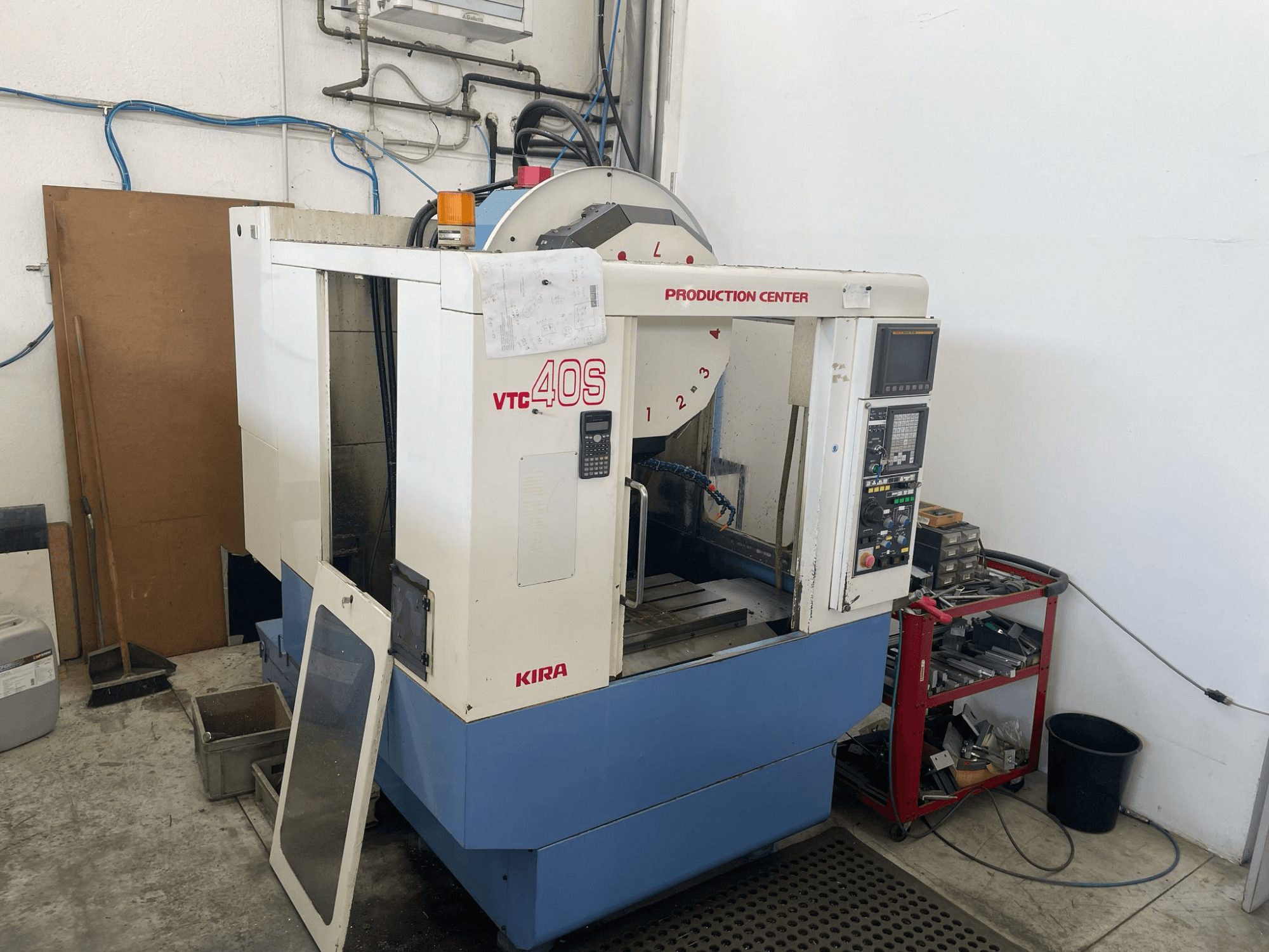 Centrum obróbcze KIRA VTC 40S w widoku z boku, z panelem sterowania, otwartymi drzwiami i narzędziami na pobliskim wózku magazynowym.
