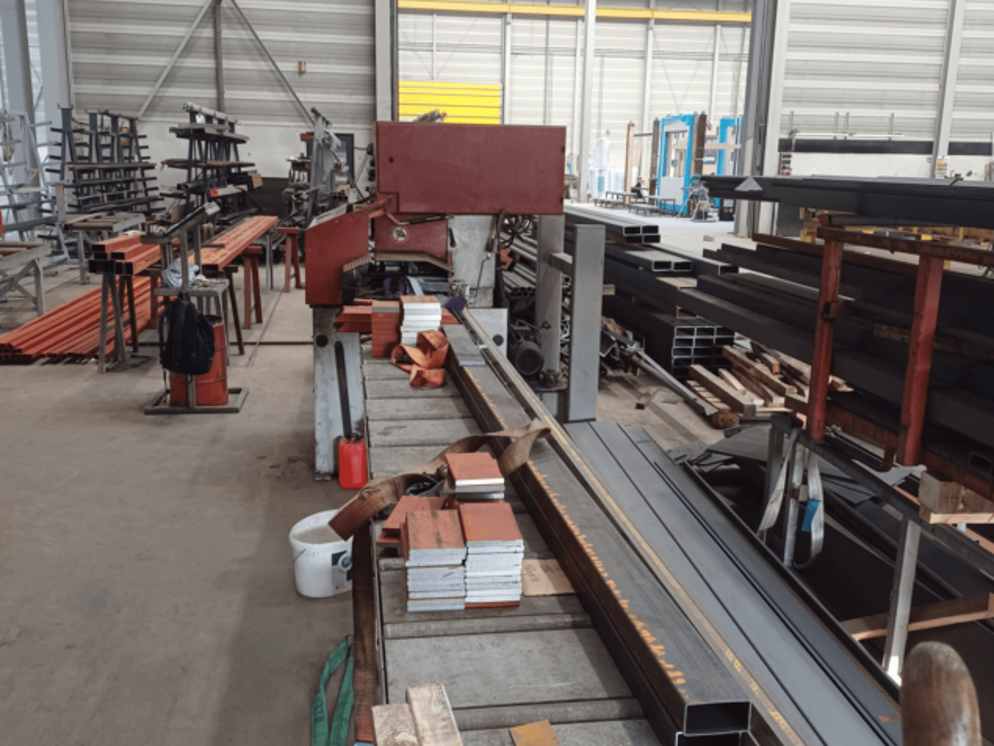 Warsztat produkcyjny z różnymi profilami metalowymi, z czerwoną piłą na pierwszym planie i ułożonymi materiałami.