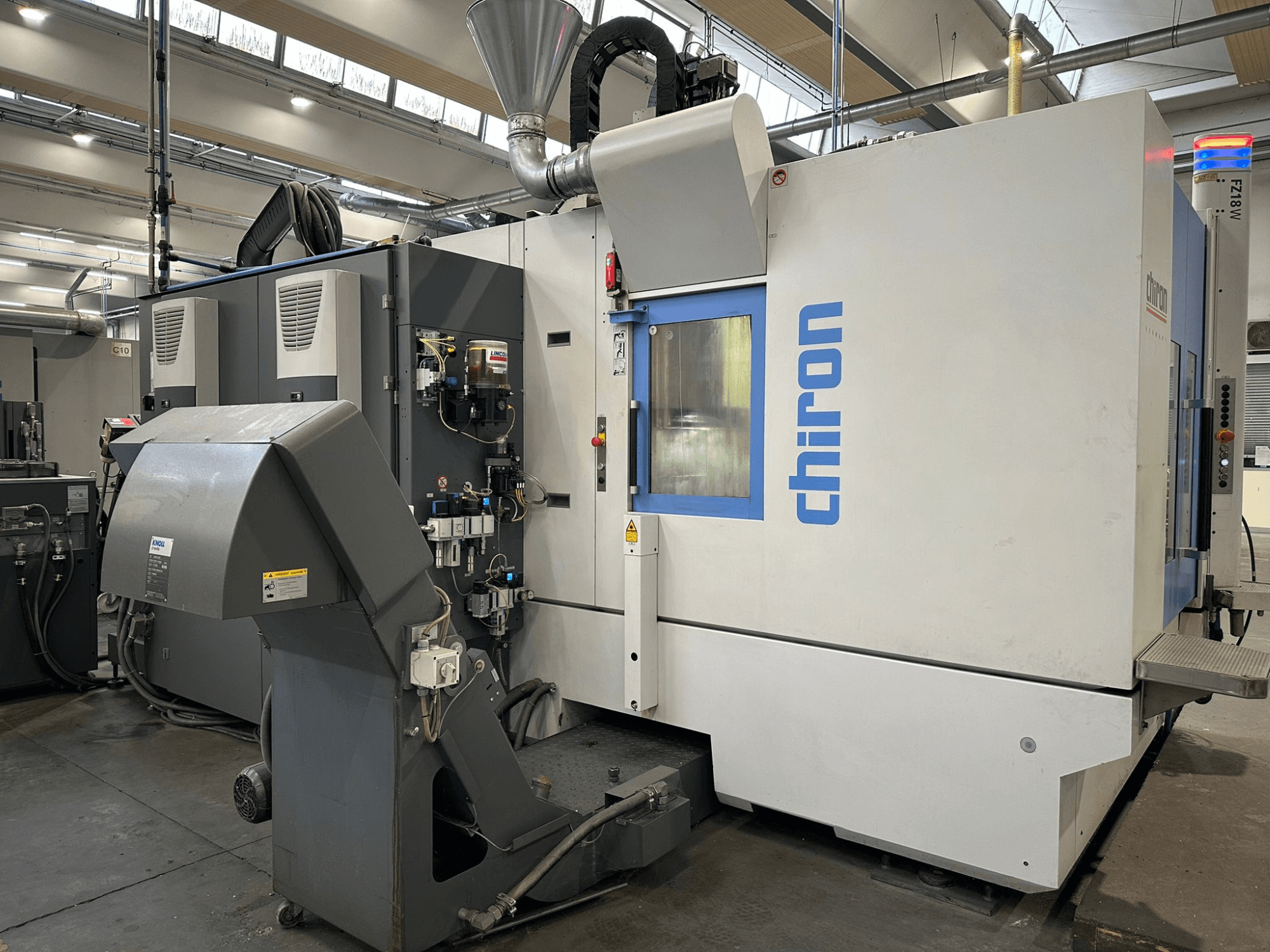 Maszyna CNC Chiron w kolorze szarym i białym, widok z przodu, z elementami sterującymi, rurami i osłoną odpylającą.