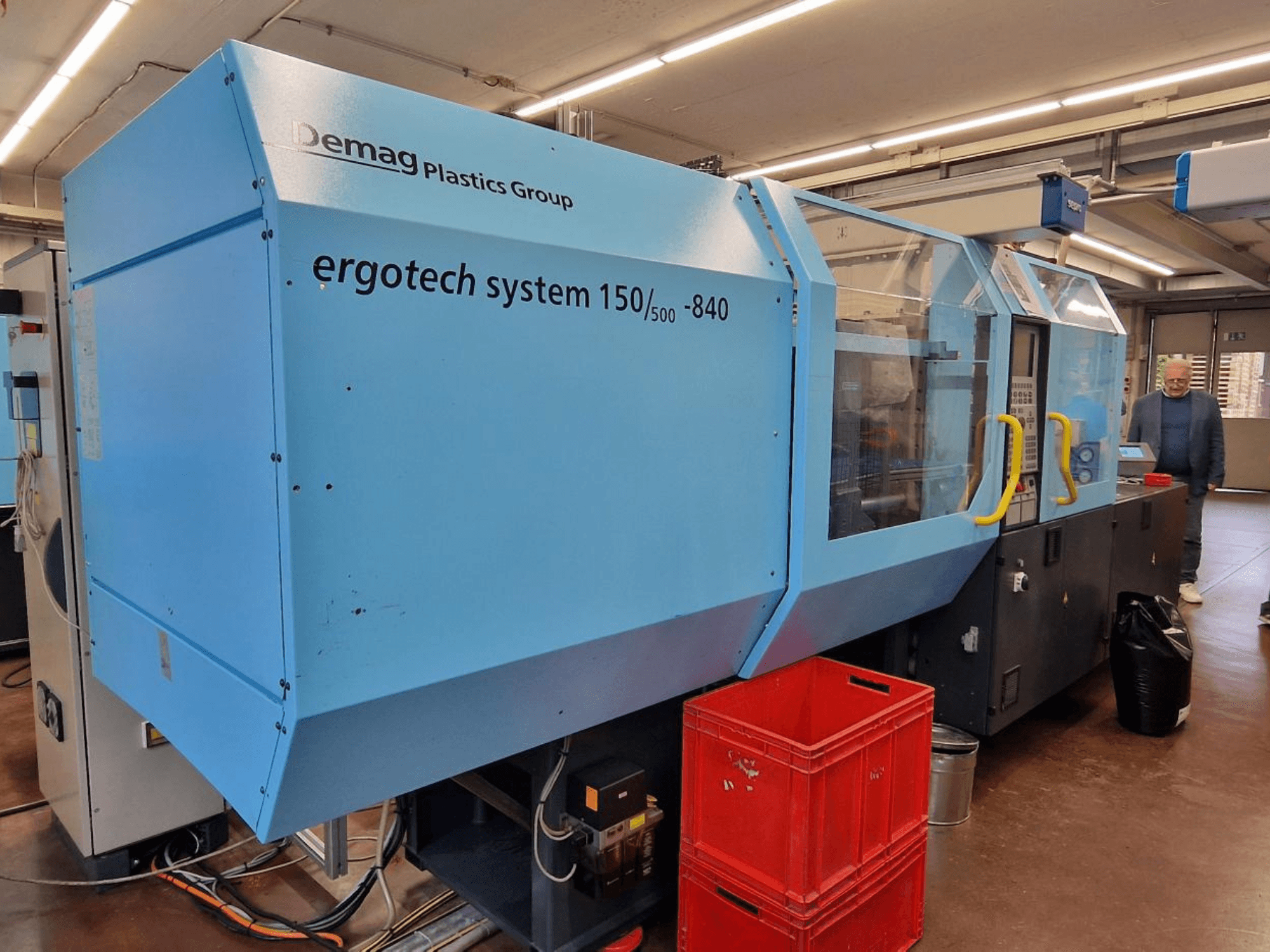 Wtryskarka Demag Ergotech System 150/500-840 w kolorze niebieskim, widok z boku, ze skrzynkami i operatorem w tle.