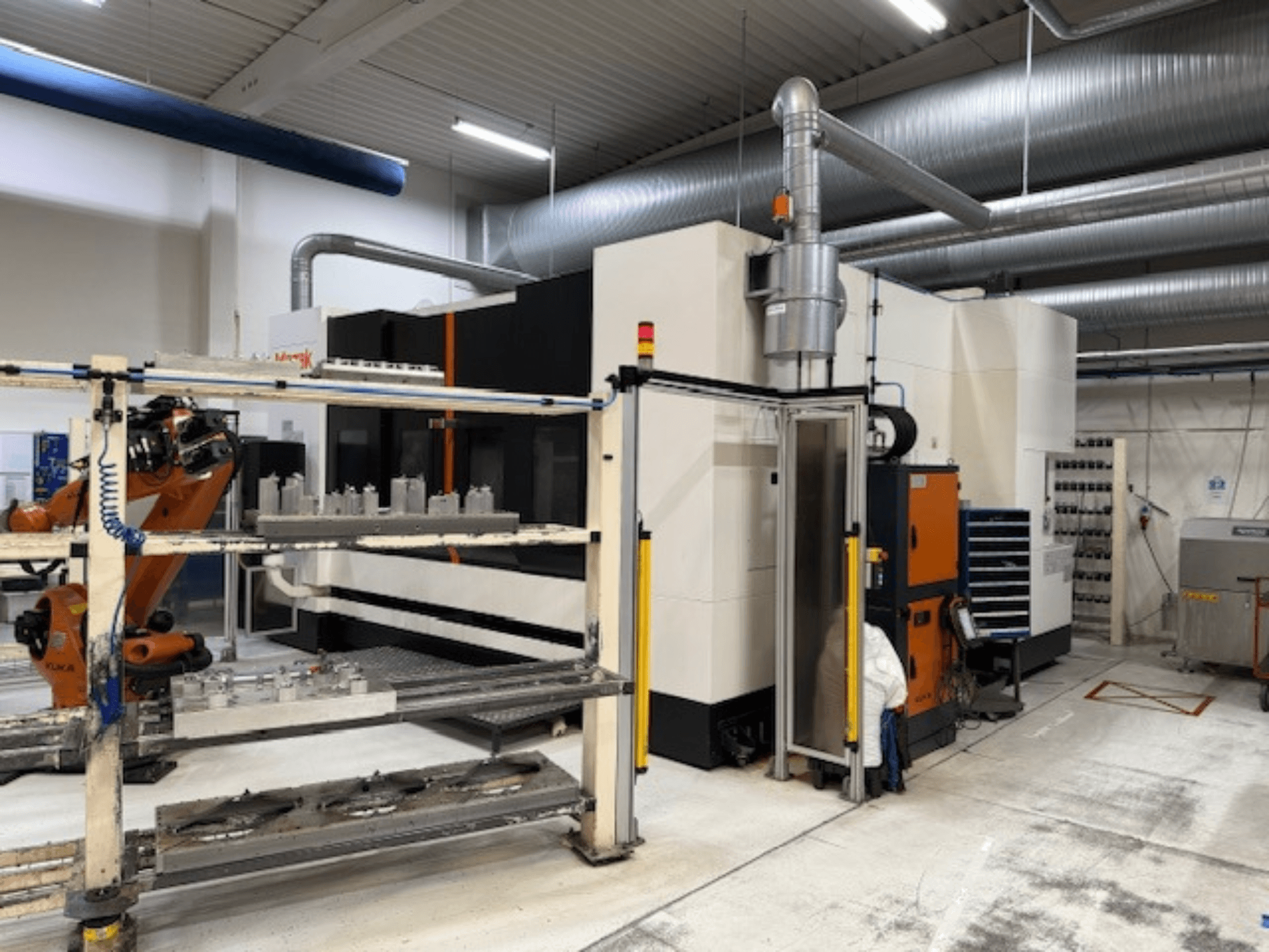 Przemysłowa maszyna CNC z ramieniem robotycznym i barierami bezpieczeństwa, z widokiem z przodu na konfigurację w przestronnej przestrzeni roboczej.