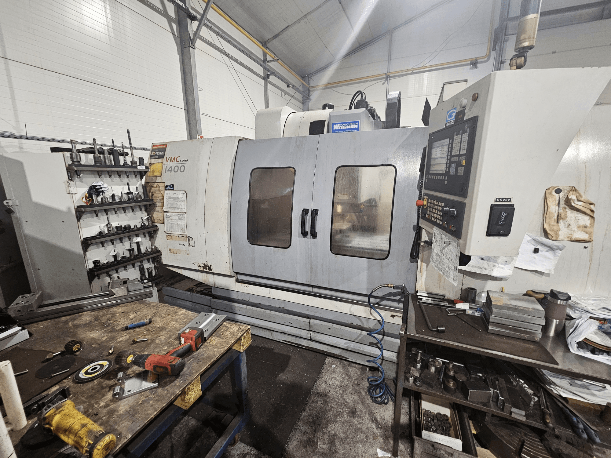 Frezarka CNC VMC 1400 w warsztacie, widok z przodu; narzędzia i materiały rozrzucone na stole warsztatowym w pobliżu.