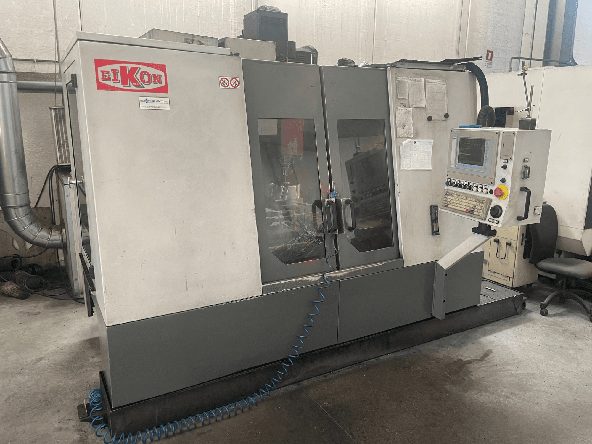 Maszyna CNC EIKON widziana od przodu, z panelem sterowania, dużymi szklanymi drzwiami i niebieskim wężem powietrza na podłodze.