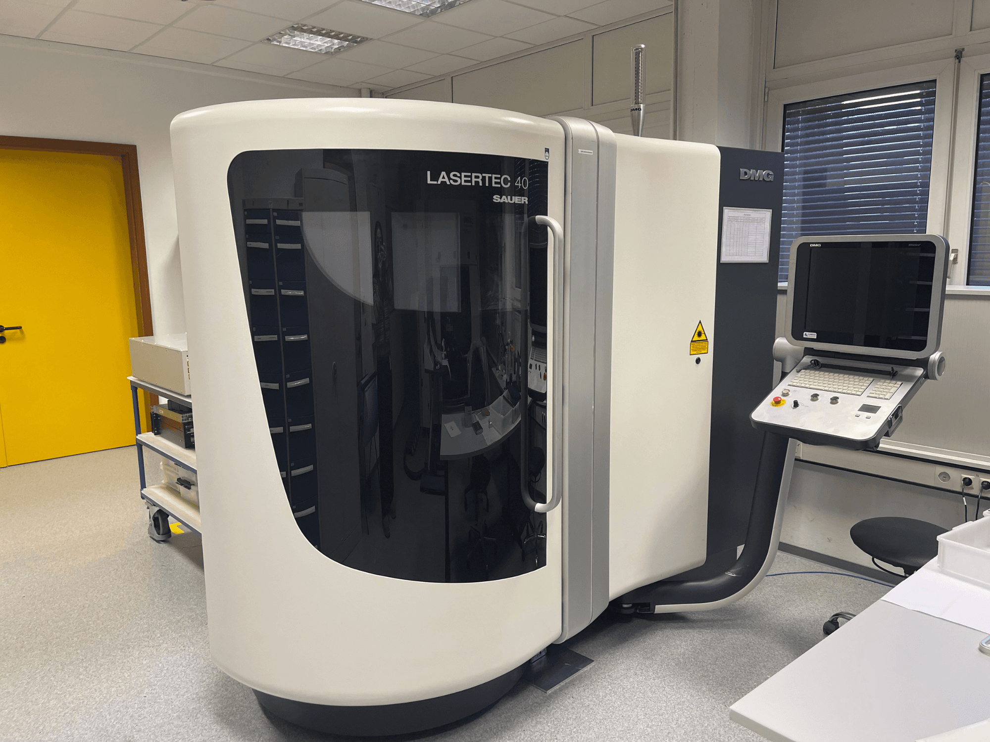 Biała frezarka LASERTEC 40 firmy DMG MORI, widok z przodu, posiada panel sterowania i ergonomiczną konstrukcję w dobrze oświetlonym pomieszczeniu.