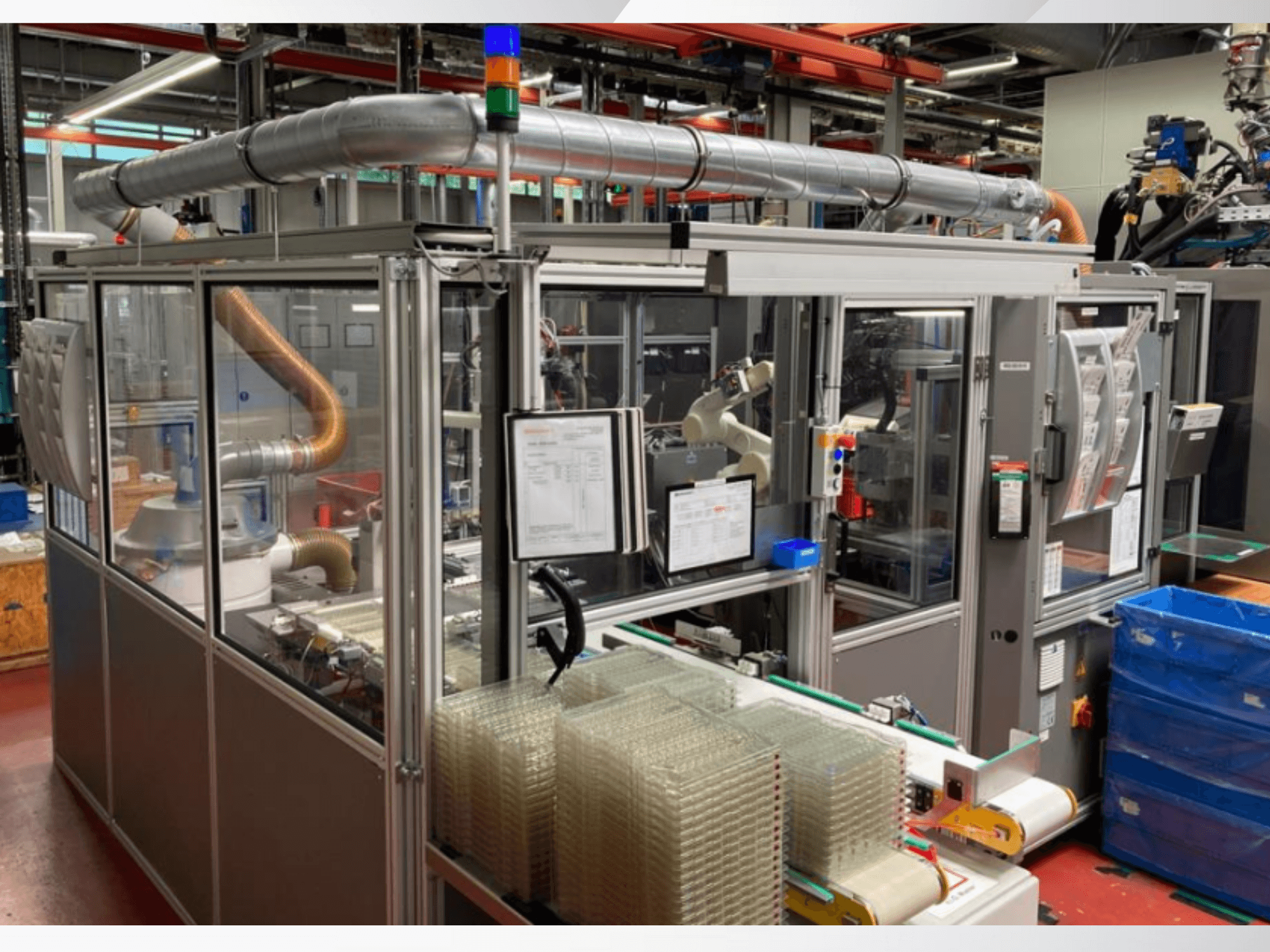 Zautomatyzowana maszyna montażowa z ramieniem robotycznym, przezroczystymi panelami bocznymi i ułożonymi w stos pojemnikami z przezroczystymi elementami, widok z przodu.