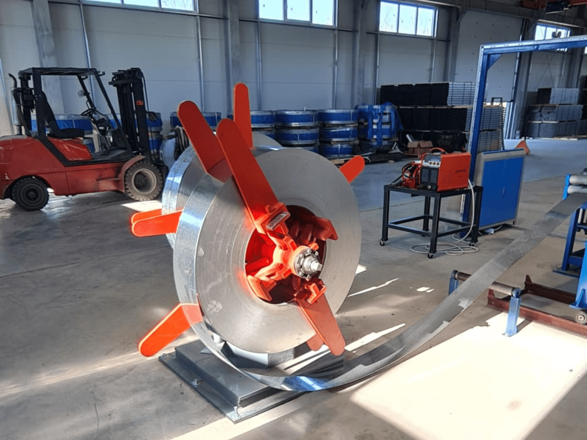 Metalowa cewka z czerwonymi uchwytami na stojaku, widziana pod kątem. Tło przemysłowe z maszynami i wózkiem widłowym.