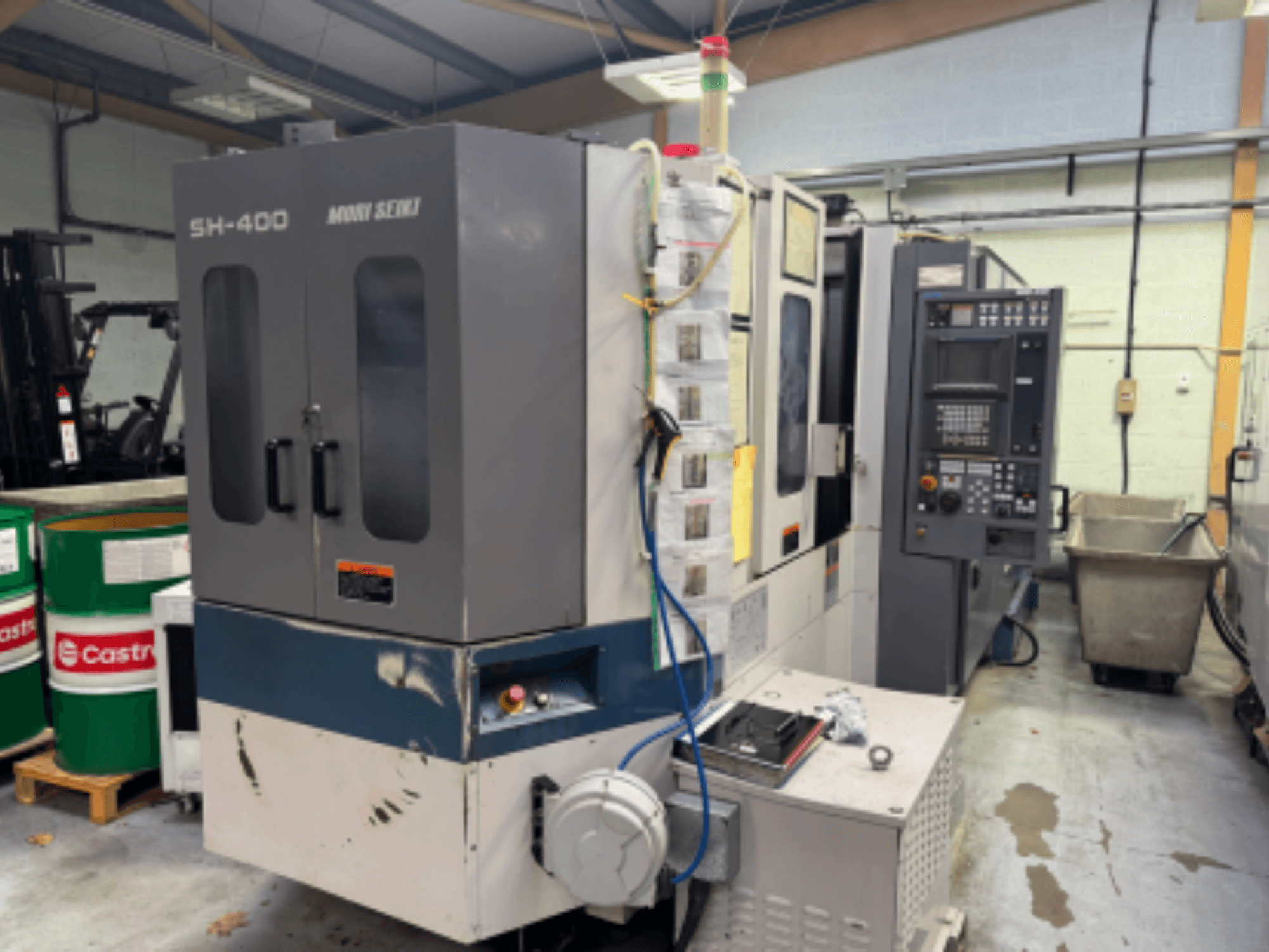 Maszyna CNC Moni Seiki SH-400, widok z boku przedstawiający panel sterowania, drzwi i otaczające środowisko przemysłowe.