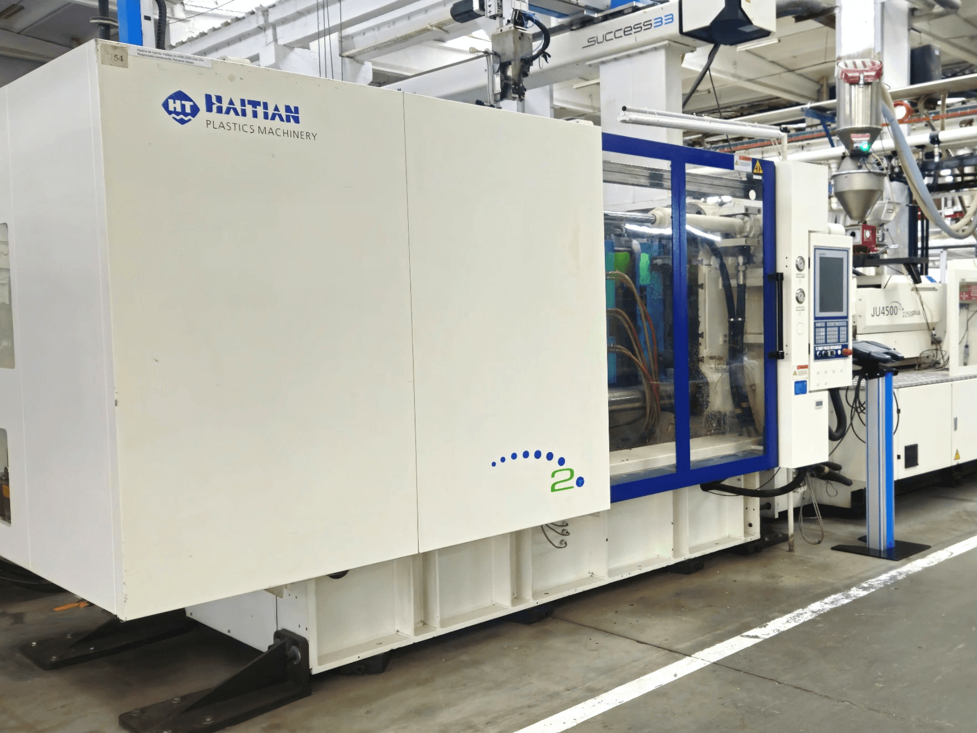 Model Haitian Plastics Machinery, widok z przodu, przedstawiający maszynę z panelem sterowania i widocznymi rurami, w otoczeniu przemysłowym.