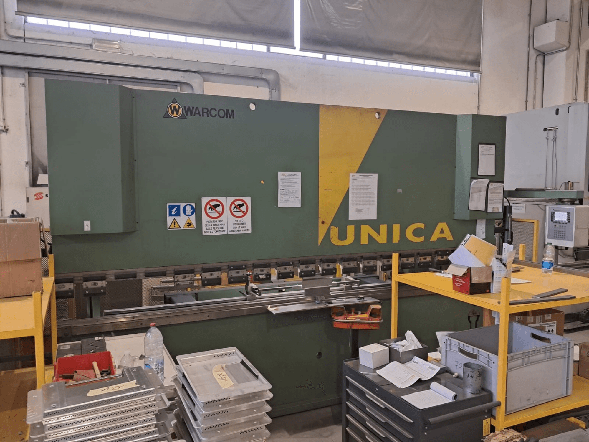 Prasa hydrauliczna Warcom UNICA 100-40, widok z przodu, zielona obudowa z żółtymi akcentami, widoczne znaki bezpieczeństwa, narzędzia i materiały na stole.