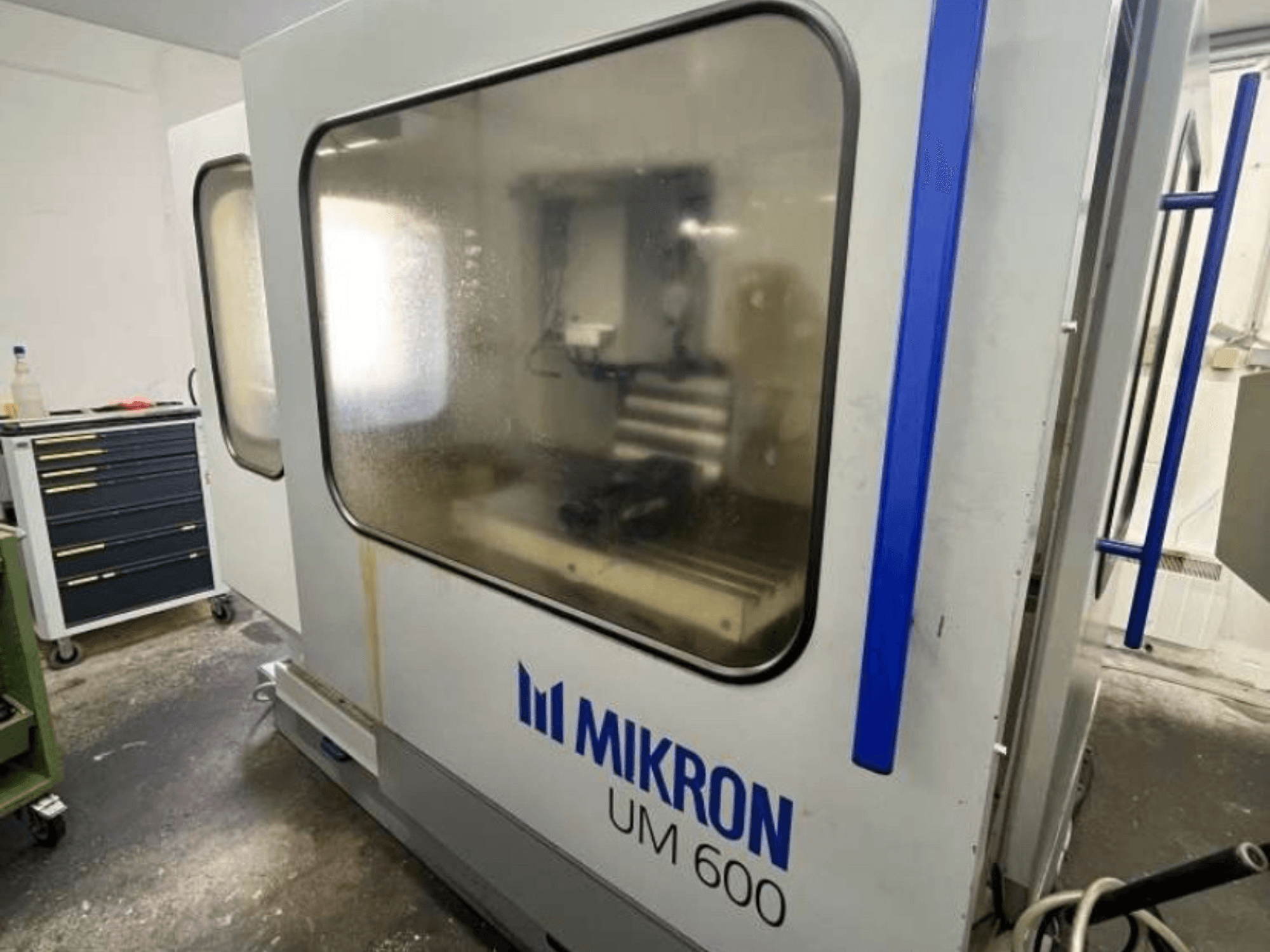 Uniwersalne centrum obróbcze MIKRON UM 600 TNC 410/426, widok z boku, z zaparowaną szybą i widocznym magazynem narzędzi w pobliżu.