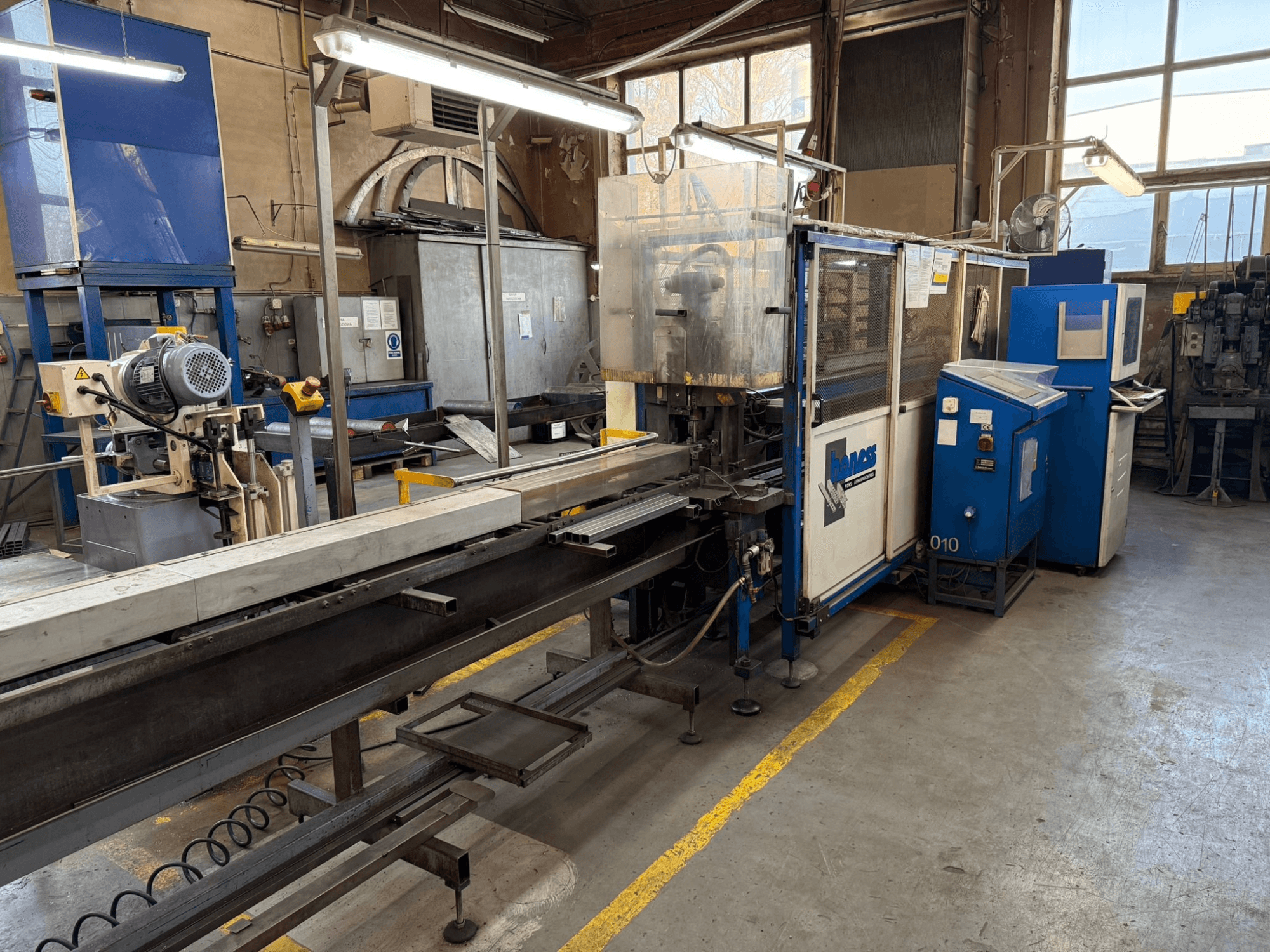 Warsztat przemysłowy z niebieską maszyną Haweas, widok z boku pokazujący przenośnik i sprzęt w dużej, dobrze oświetlonej przestrzeni.