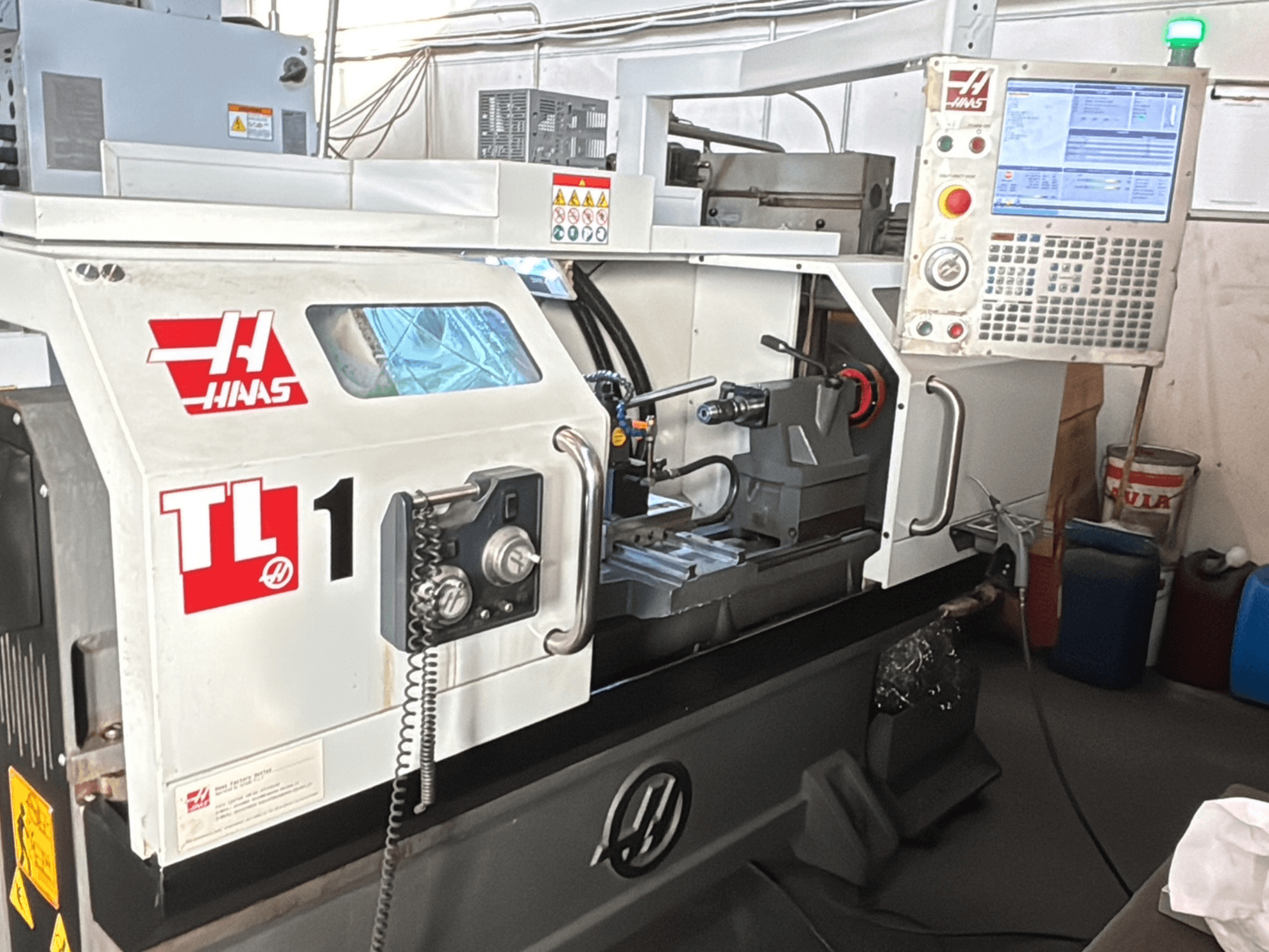 Tokarka CNC Haas TL-1, widok z przodu, posiada panel sterowania z wyświetlaczem, uchwyt narzędziowy i etykiety bezpieczeństwa.