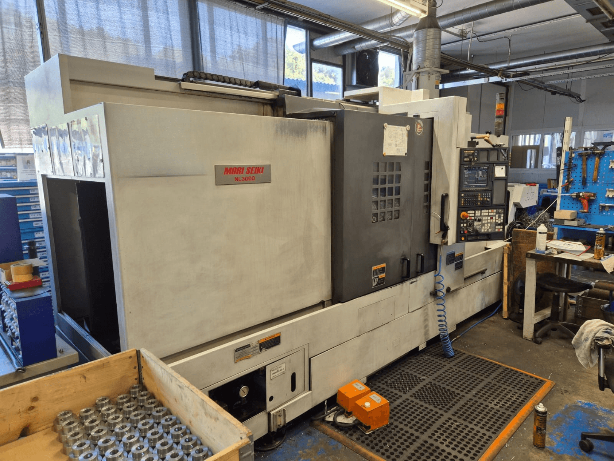 Tokarka CNC Mori Seiki NL3000 w warsztacie, widok z boku z panelem sterowania, narzędziami i metalowymi częściami w drewnianej skrzynce.