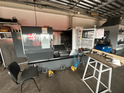 Tokarka CNC Haas (model ST-10) w warsztacie, widok z boku przedstawiający elementy sterujące, narzędzia na stole warsztatowym i krzesło w pobliżu.
