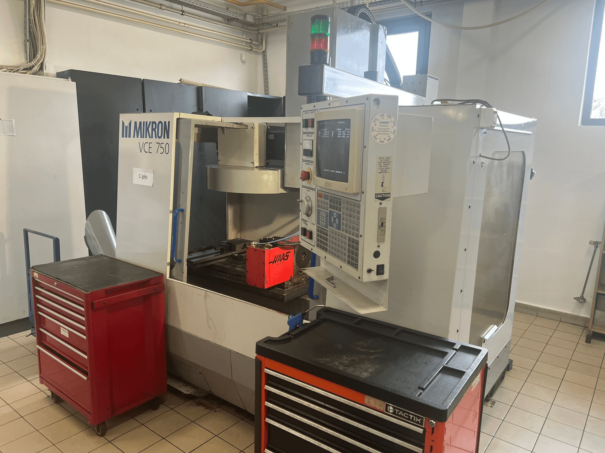 Alt text: "Pionowe centrum obróbcze MIKRON VCE 750, widok z przodu z panelem sterowania i czerwonymi skrzynkami narzędziowymi, w przestronnym warsztacie".