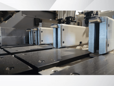 Widok z bliska mechanizmu zaciskowego maszyny precyzyjnej z metalowymi szczękami, z czarnymi powierzchniami roboczymi i widocznymi prowadnicami.