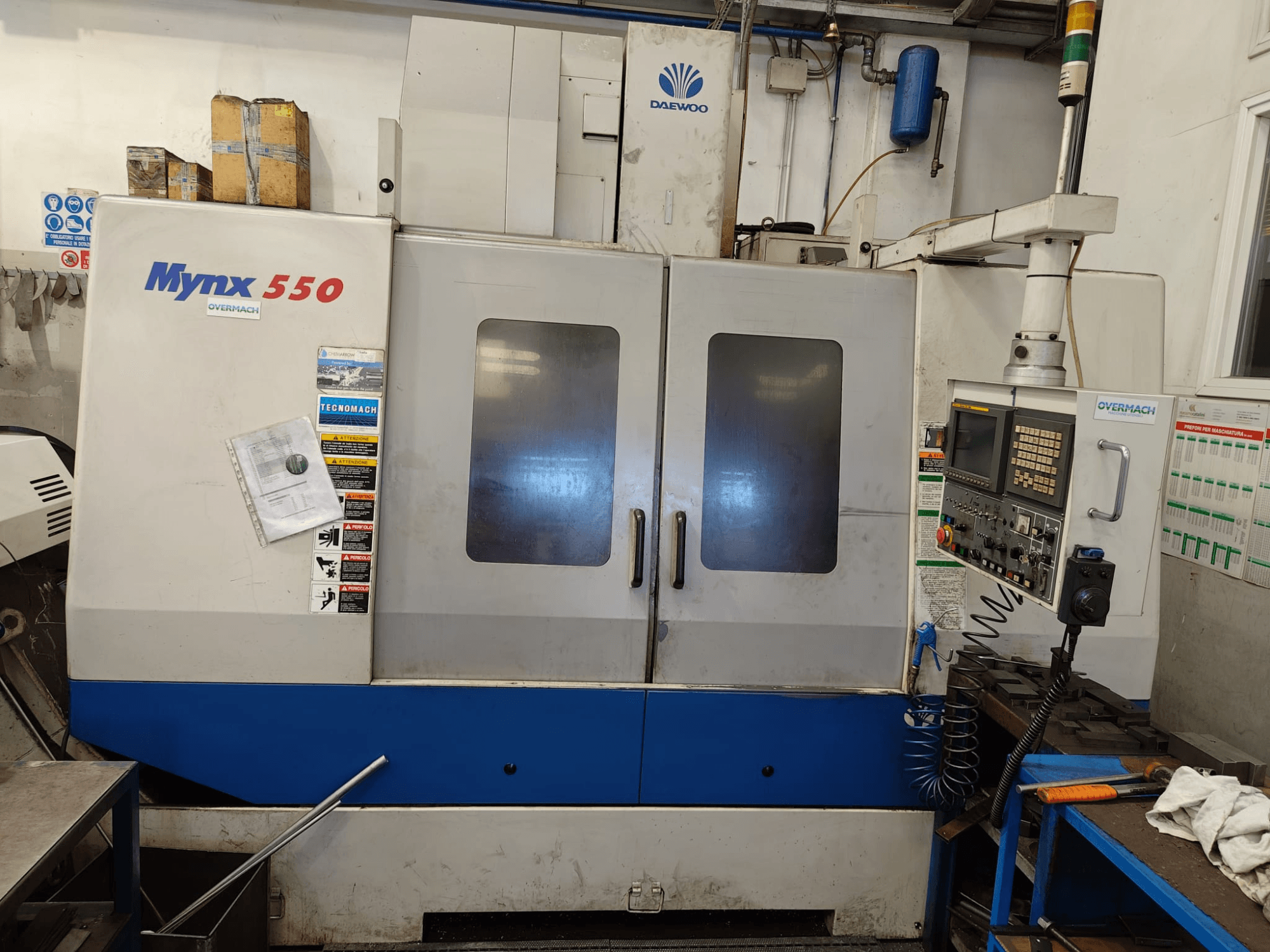 Pionowe centrum obróbcze DAEWOO MYNX 550, widok z przodu, z panelem sterowania, dwojgiem dużych drzwi i etykietami bezpieczeństwa.