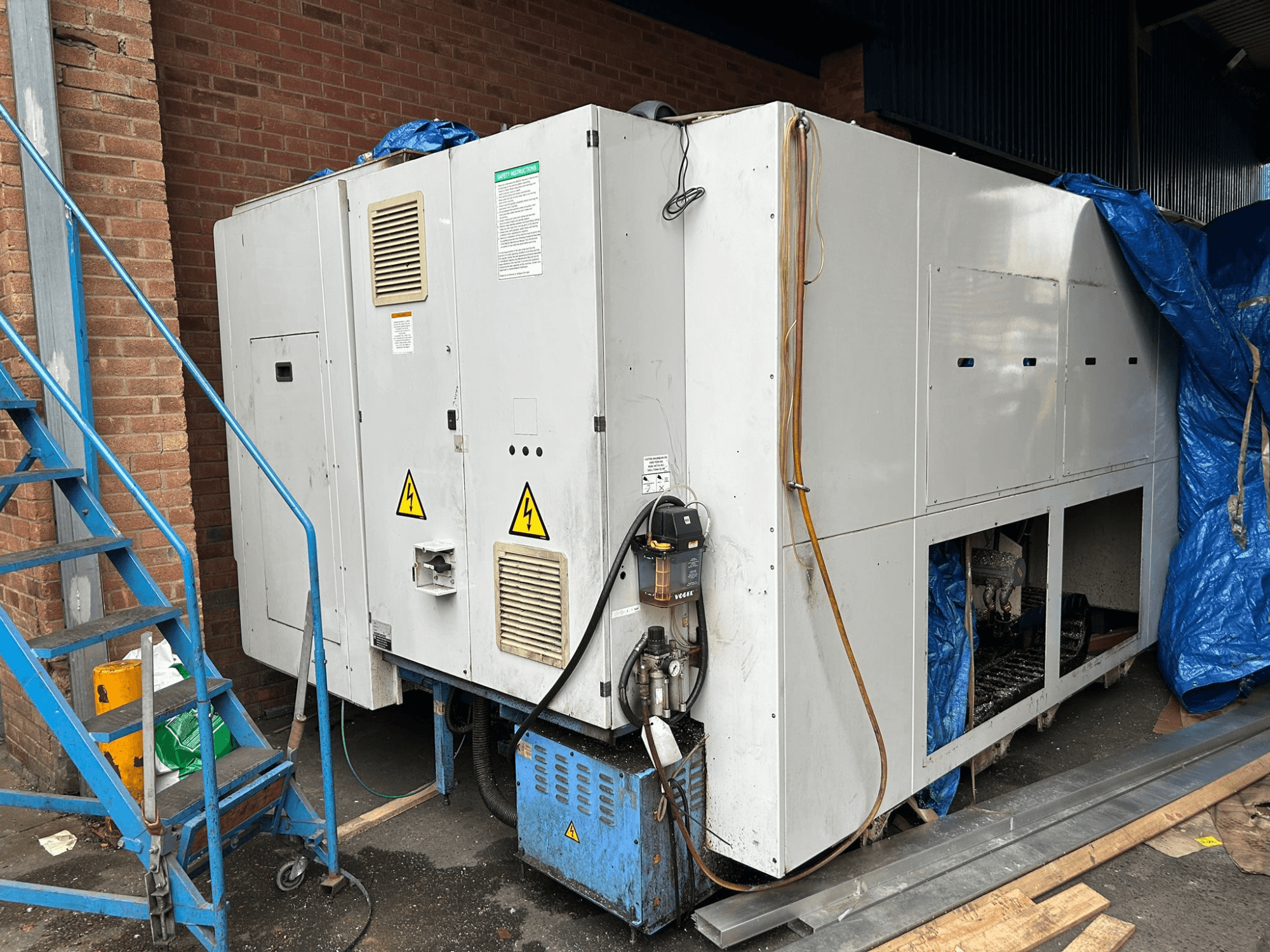 Widok z boku dużego przemysłowego agregatu chłodniczego, marka i model niejasne, pokazujący połączenia elektryczne, znaki ostrzegawcze i niebieską plandekę.