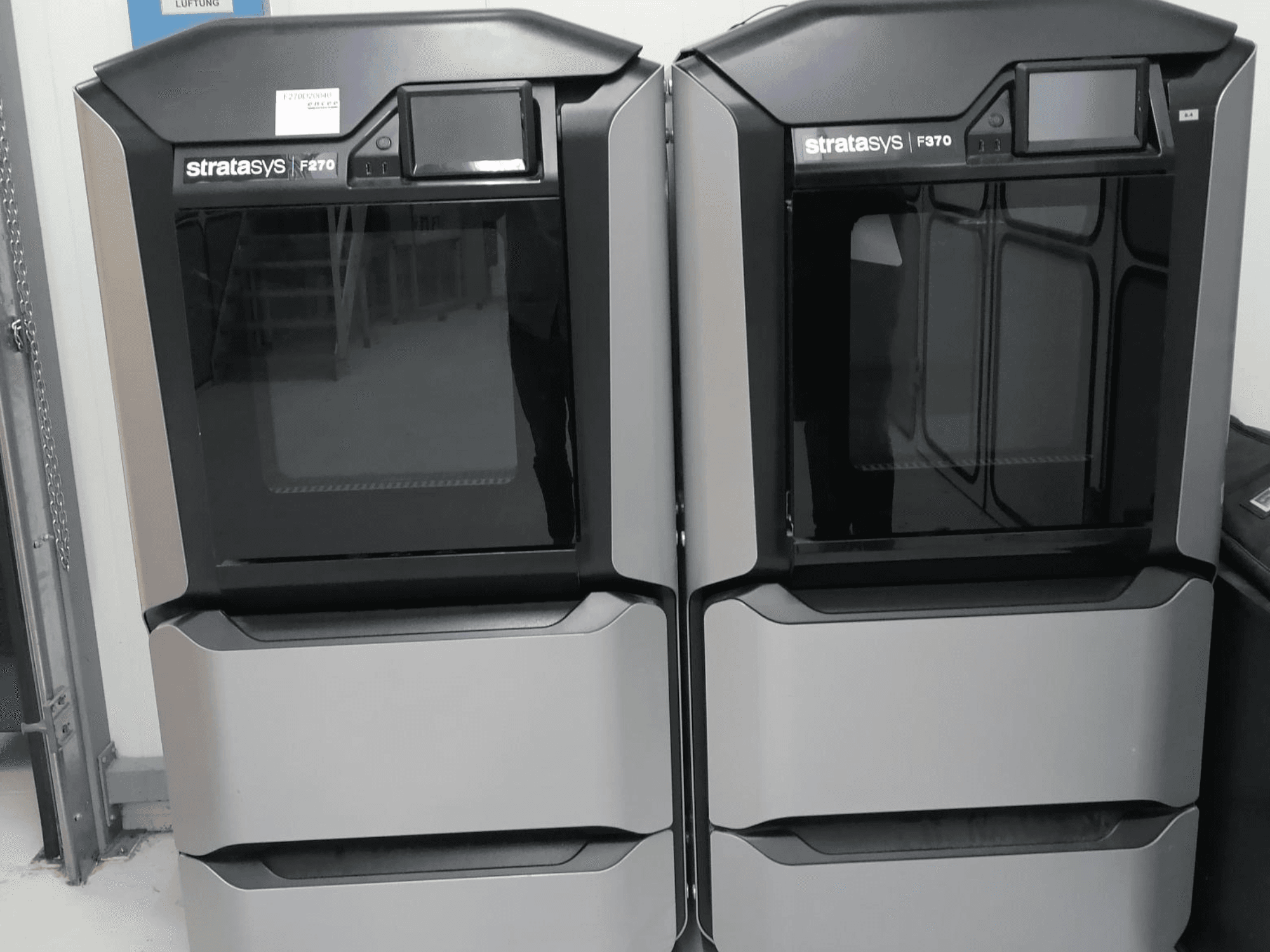 Drukarka 3D do tworzyw sztucznych Stratasys F270 z widokiem z przodu, wyposażona w dwie maszyny z eleganckimi szarymi obudowami i cyfrowymi wyświetlaczami.
