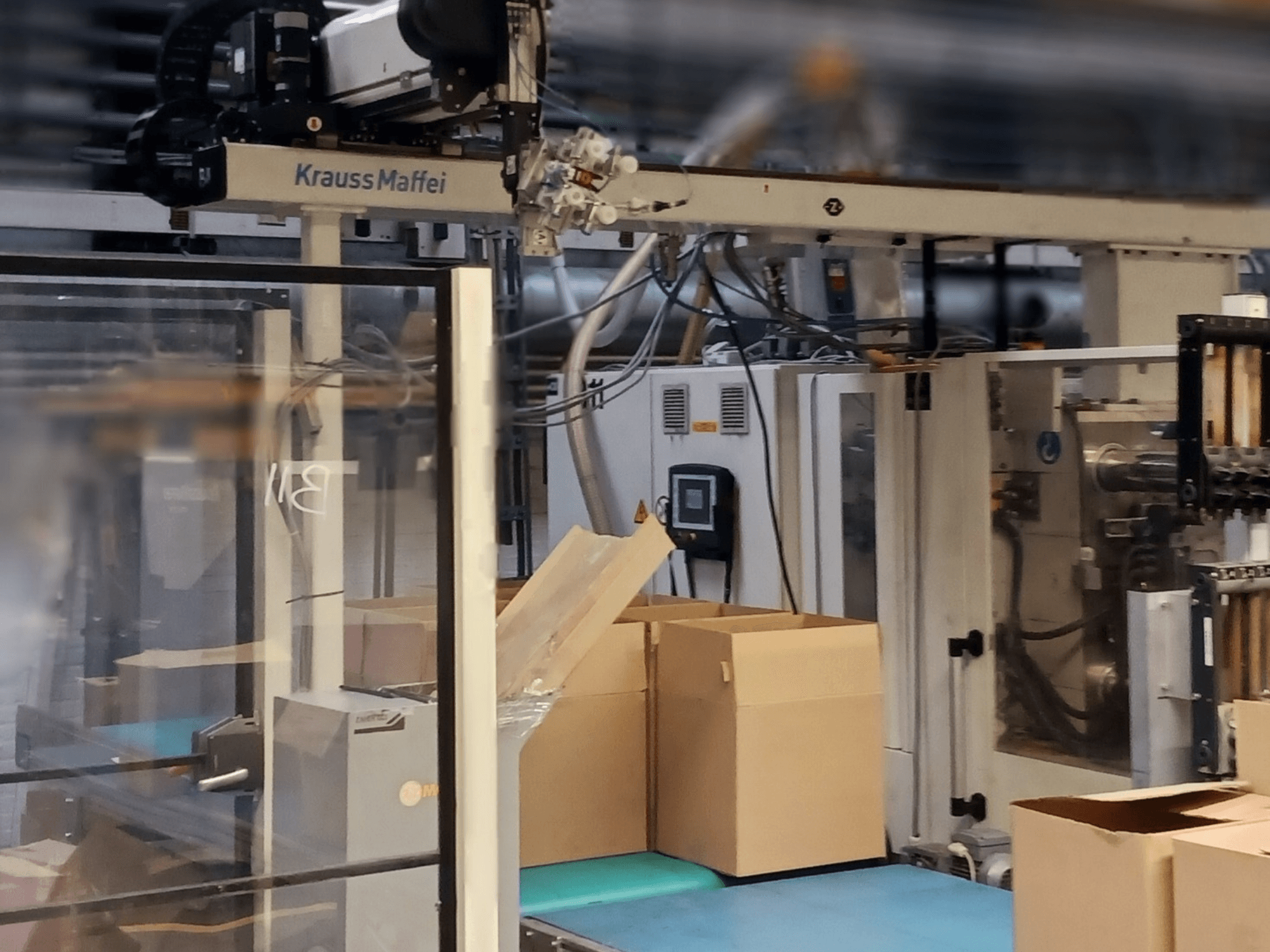 System automatyki Krauss Maffei w fabryce, widok z przodu przedstawiający skrzynki na przenośniku i działające komponenty maszyn.
