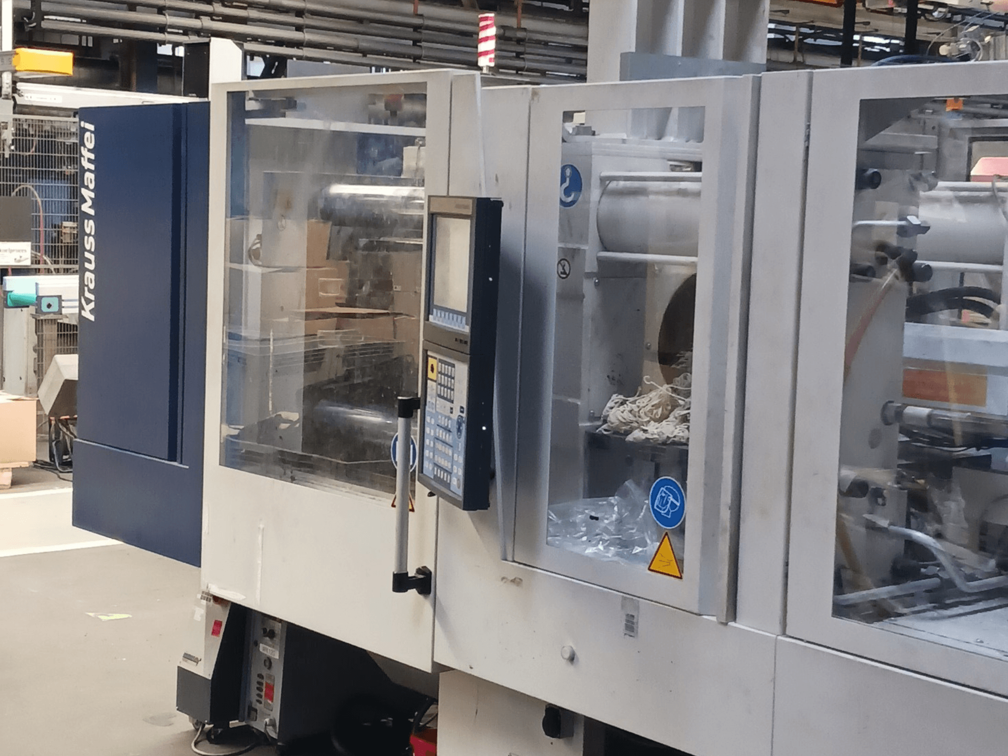 Wtryskarka KraussMaffei, widok z boku, pokazujący panel sterowania, maszyny i materiały opakowaniowe w tle.