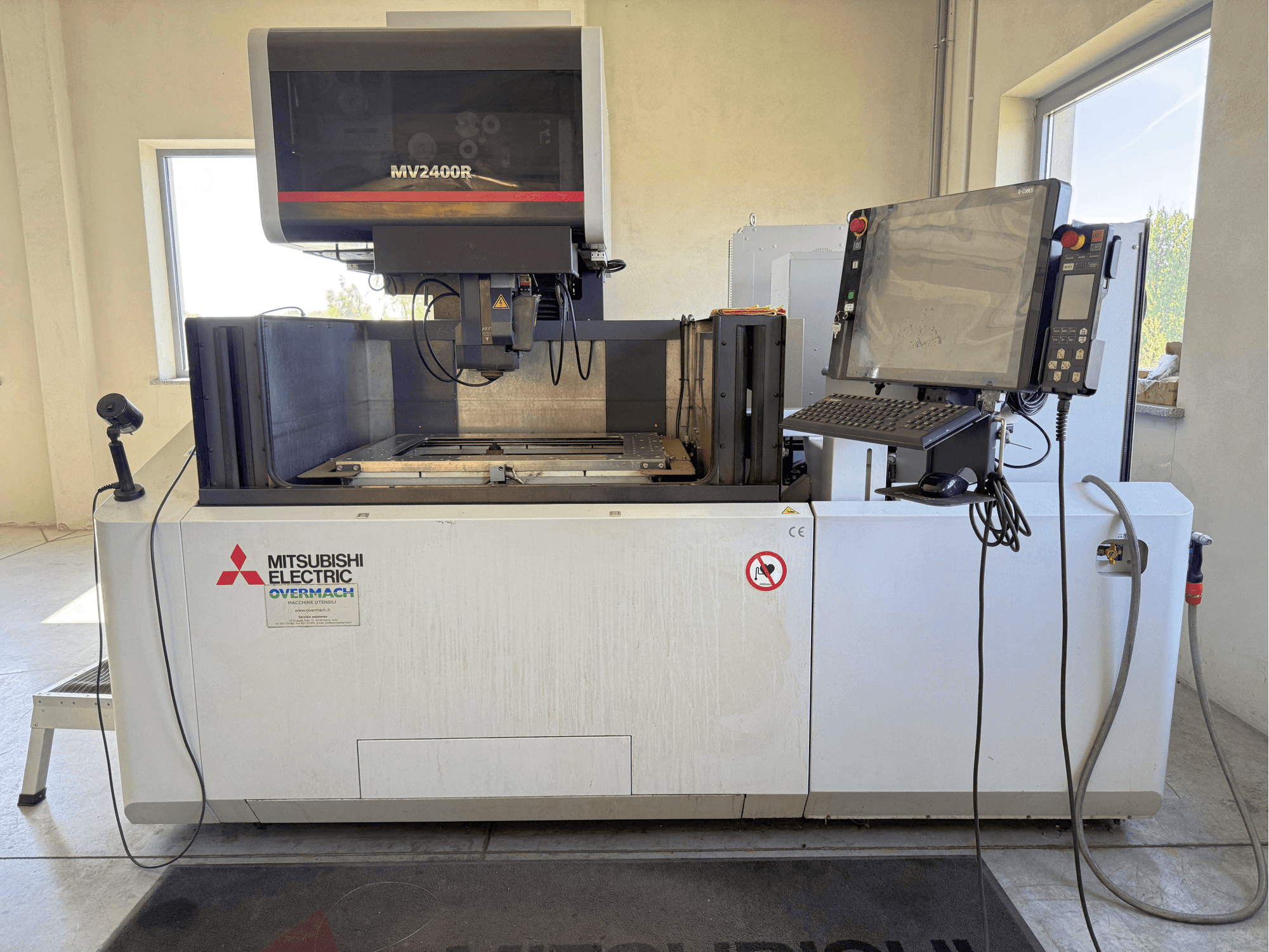 Maszyna do elektroerozyjnego cięcia drutowego Mitsubishi MV2400R – widok z przodu, przedstawiający panel sterowania, obszar roboczy oraz oznakowanie bezpieczeństwa.