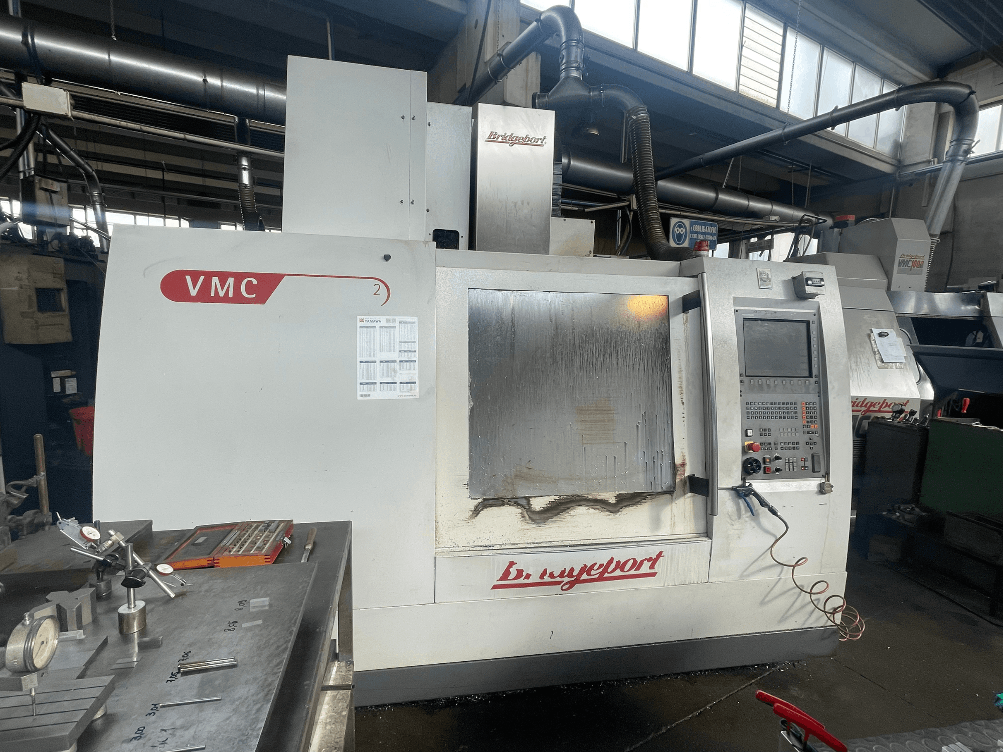 Pionowe centrum obróbcze Bridgeport VCM 800-30 w warsztacie, wyposażone w panel sterowania i stół narzędziowy. Widok z przodu z widocznymi szczegółami.