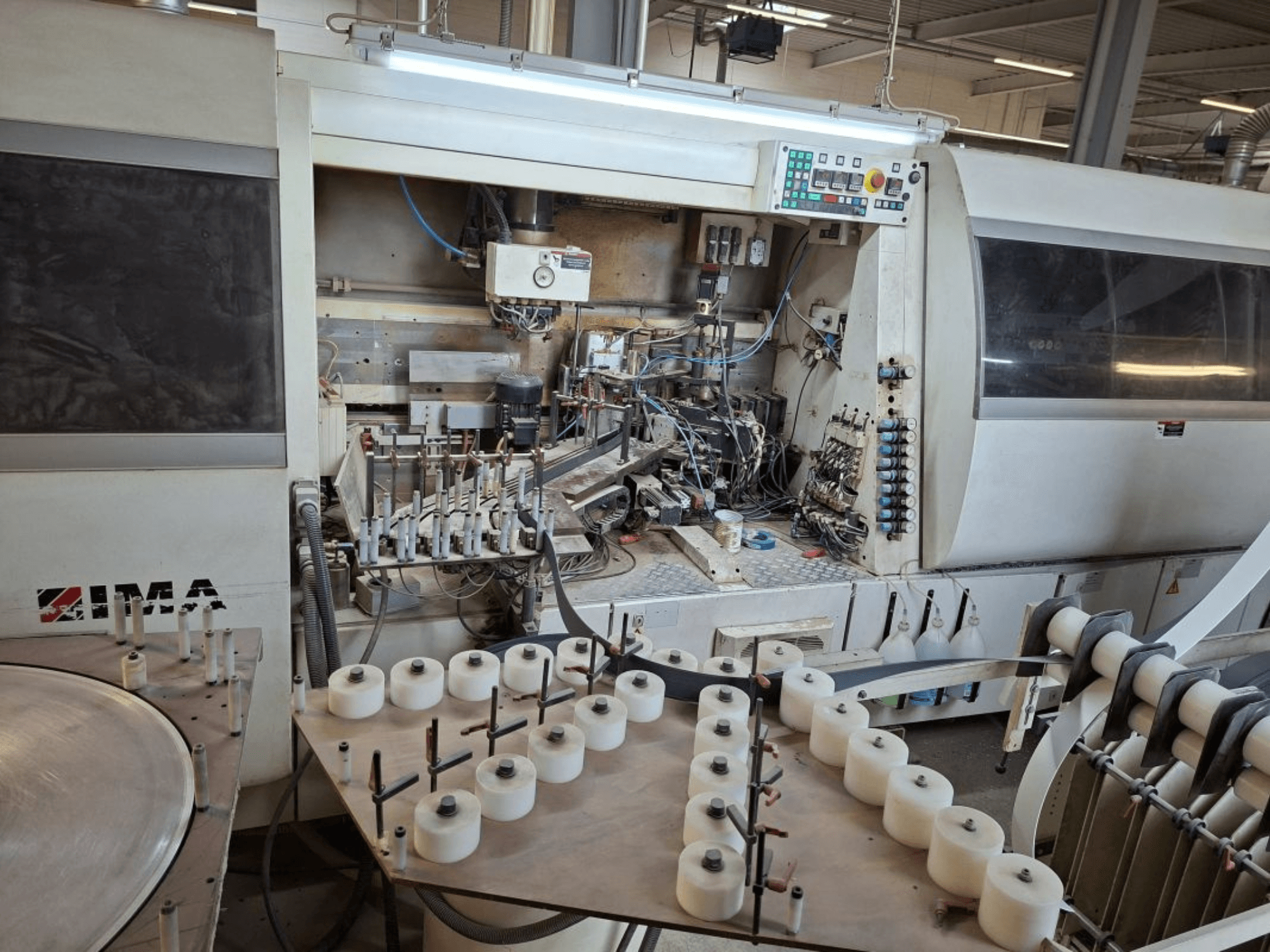 Maszyny IMA widziane z boku, prezentujące złożone komponenty mechaniczne i okrągły stół z białymi szpulami.