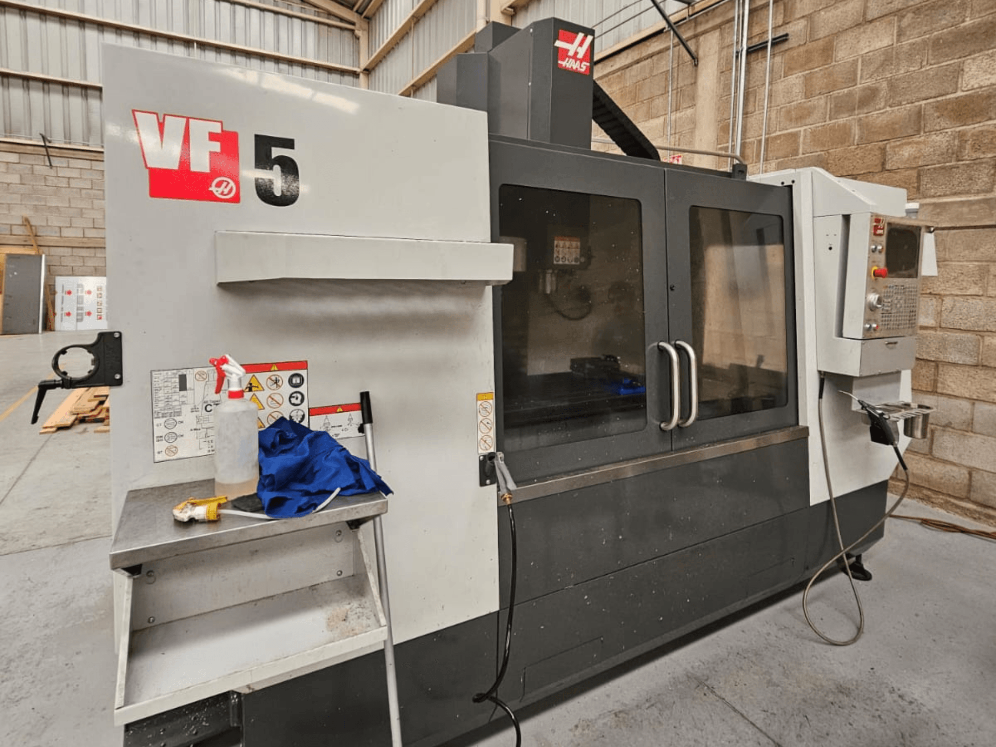 Frezarka CNC Haas VF-5 w widoku z przodu, z panelem sterowania, butelką chłodziwa, niebieską ściereczką i instrukcjami bezpieczeństwa.