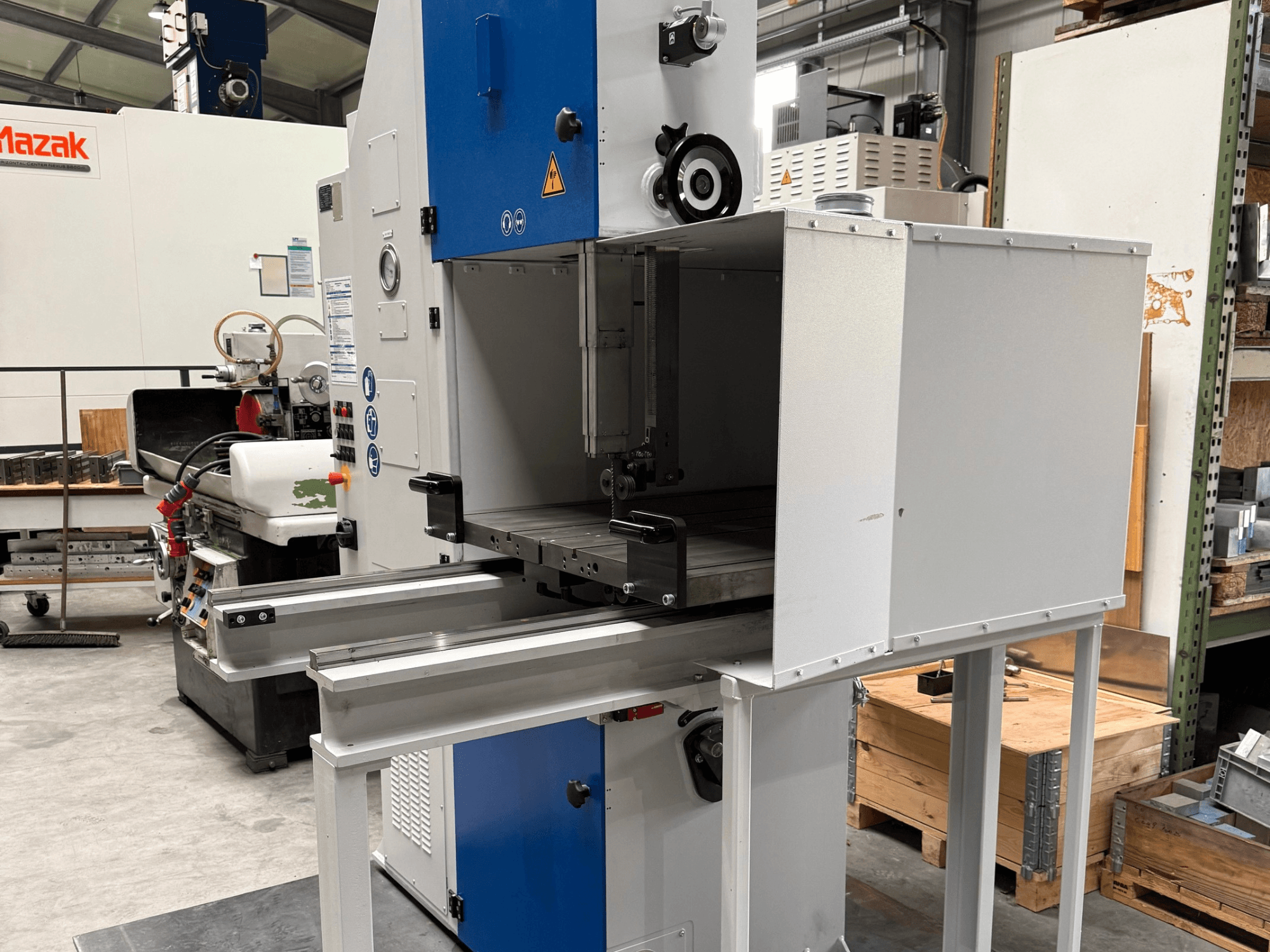 Maszyna przemysłowa Mazak widziana od przodu, z niebieskim panelem sterowania, metalowym stołem i ostrzeżeniami dotyczącymi bezpieczeństwa.