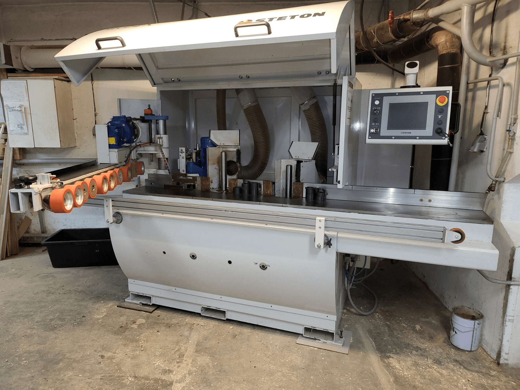 Widok z przodu maszyny do obróbki drewna firmy Stetton z panelem sterowania, rurami podajnika i różnymi kawałkami drewna na stole.