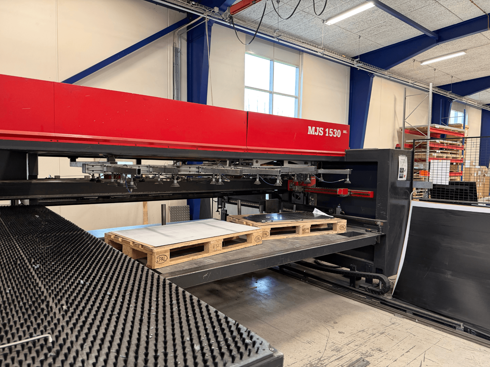 Maszyna CNC MJS 1530 w warsztacie, widok z przodu, z drewnianą paletą trzymającą arkusz metalu na podniesionej platformie.
