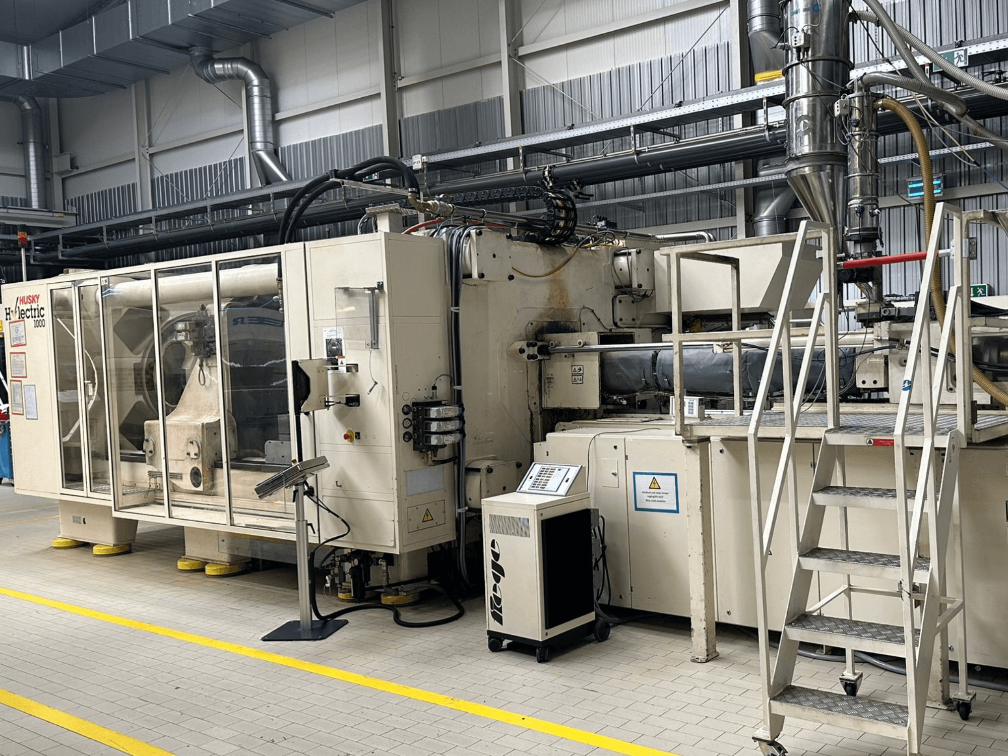 Widok z przodu elektrycznej wtryskarki Husky Hylelectric H1000 RS95/80 w warunkach fabrycznych, z panelem sterowania i schodkami.