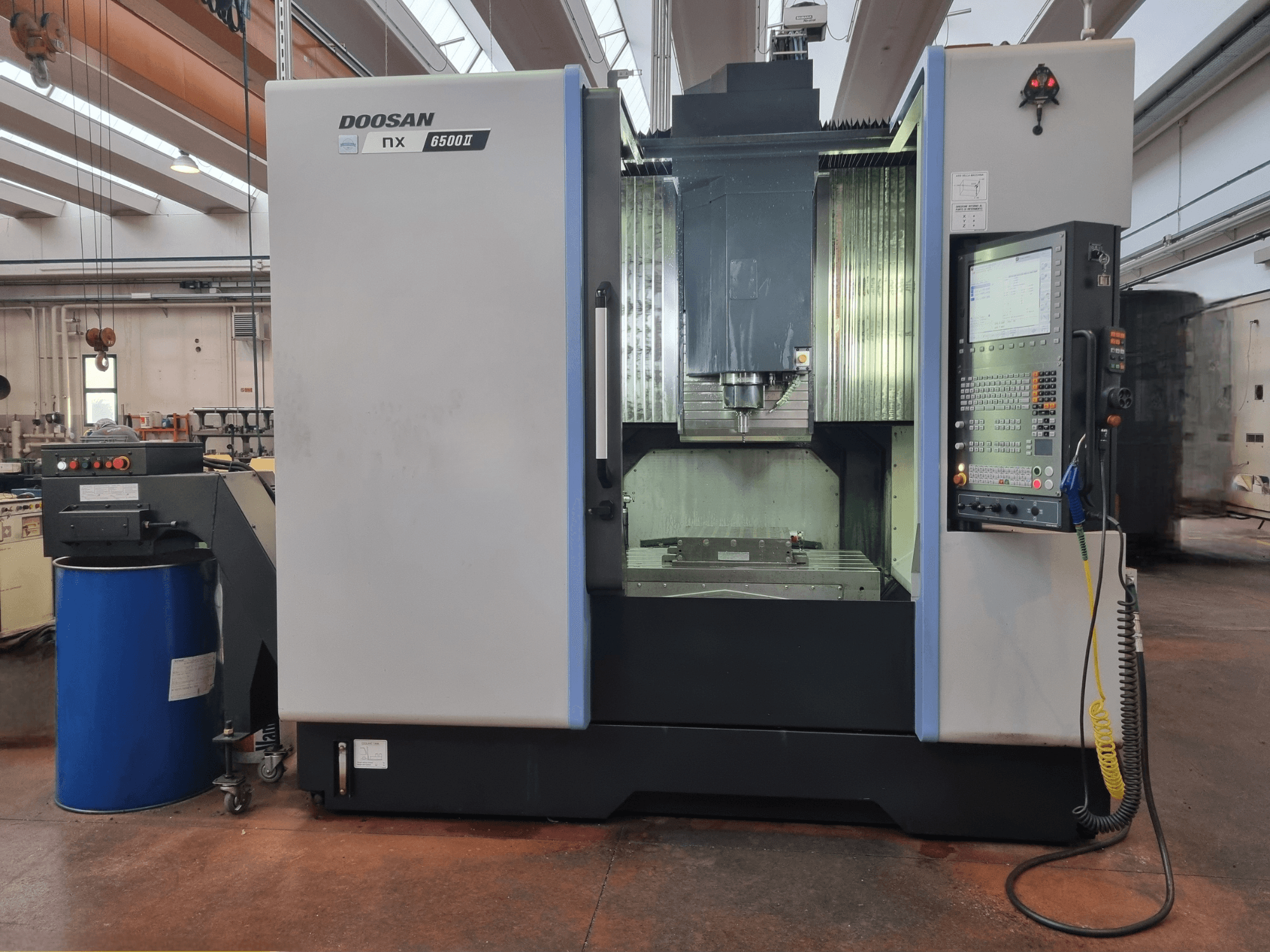 Maszyna CNC Doosan NX 6500 II, widok z przodu, z panelem sterowania i obszarem obróbki, umieszczona obok niebieskiej beczki.
