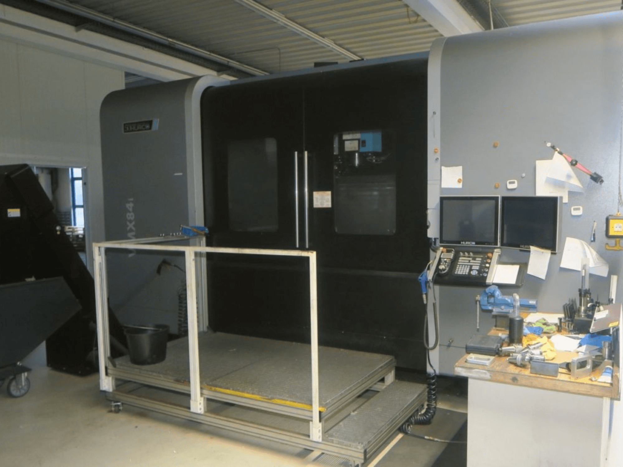 Maszyna CNC Hurco MX84 w widoku z przodu, pokazująca dwa monitory, panel sterowania i obszar roboczy z różnymi narzędziami.