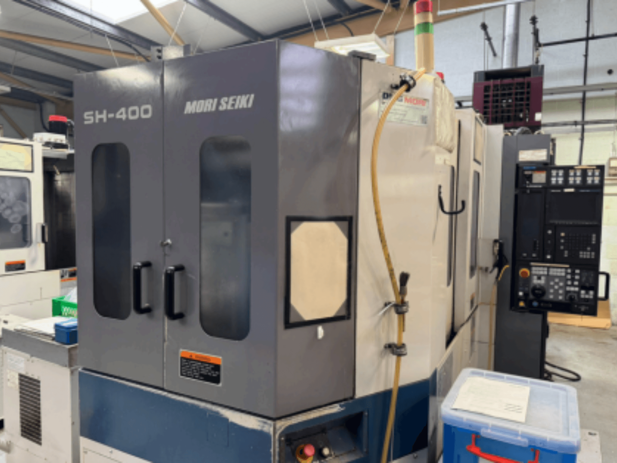 Maszyna CNC Mori Seiki SH-400, widok z przodu, z podwójnymi drzwiami, panelem sterowania i wężem mocującym. Niebieski pojemnik do przechowywania w pobliżu.