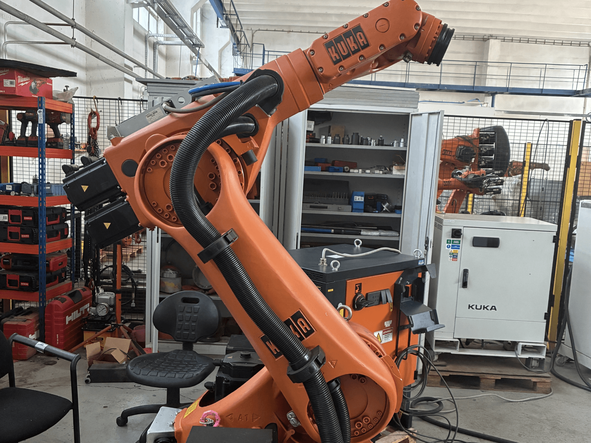 Ramię robota KUKA KR30-3 w środowisku przemysłowym, widok z boku pokazujący kable i elementy sterujące, z narzędziami i półkami w tle.